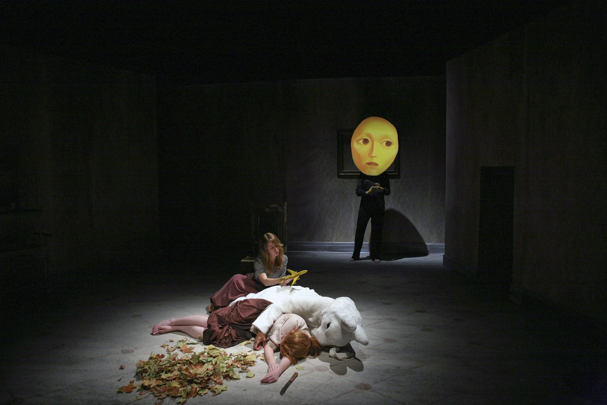 The image depicts a scene from a theatrical performance showing children on stage. Some children are lying on the floor, covered with costumes resembling animals or characters, while others are sitting. In the background, a performer with a large, ro