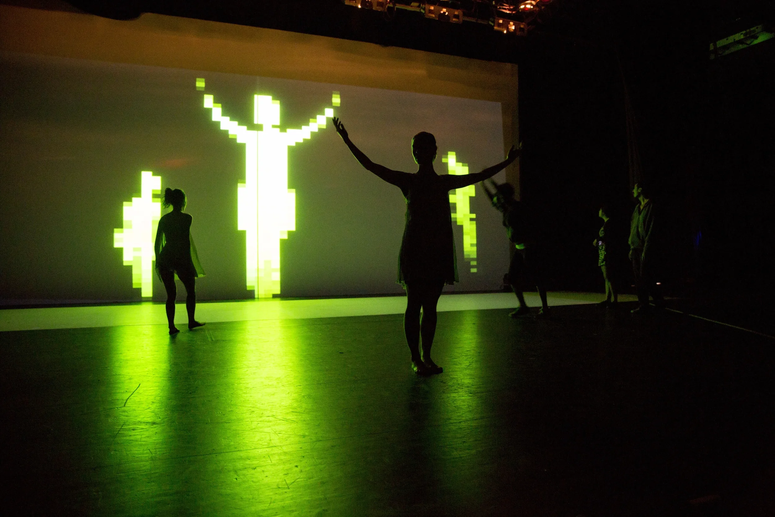 Silhouetted dancers performing in front of a large digital pixelated deer graphic projected onto a wall, with a green glow on the dance floor.