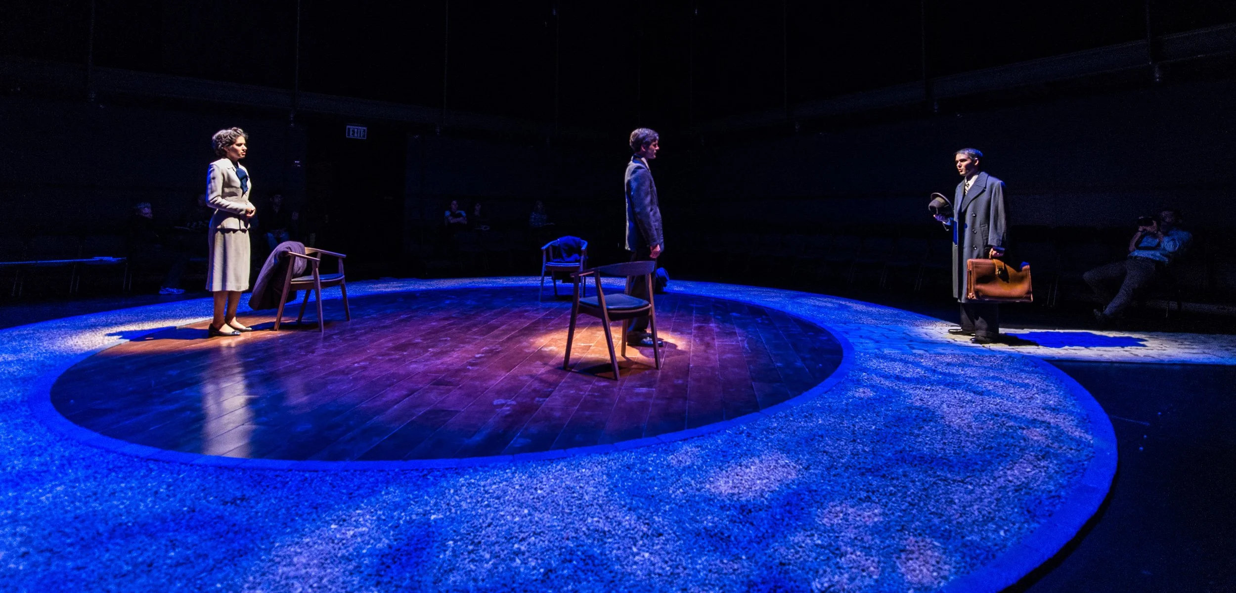 The image features a theater stage with three actors - two women and one man - engaged in a scene. The stage is circular with a wooden floor and is illuminated with blue lighting. There are chairs on the stage, and the background is dark with only a 