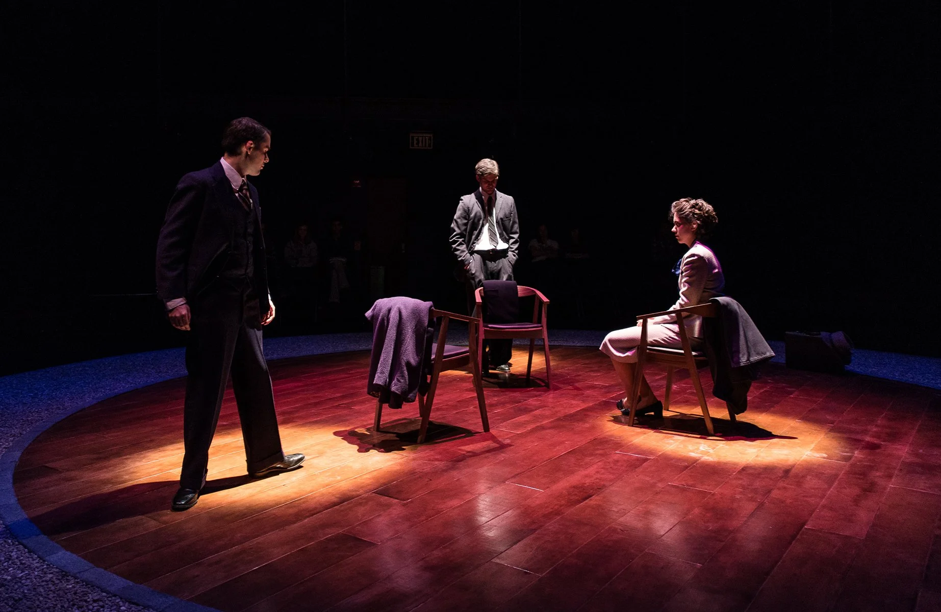 Three actors performing a scene on a dimly lit stage with wooden flooring, two are seated at a table, and one is standing, dressed in vintage clothing.