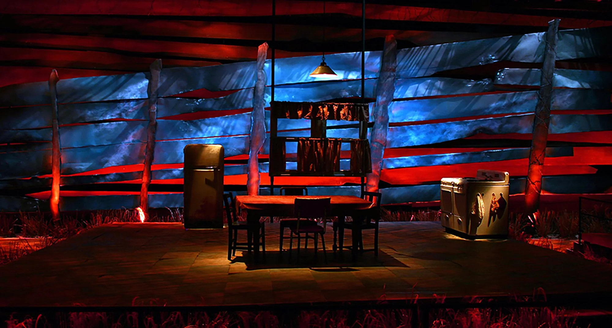 A rustic outdoor scene at night with a wooden table and chairs, illuminated by red and blue lighting, with a decorative wooden fence, a refrigerator, and a vintage-style cooler.