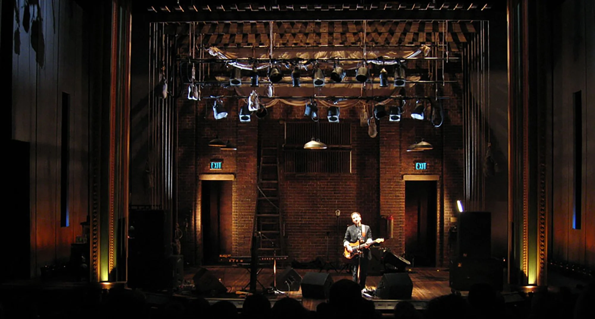 A musician playing electric guitar on stage in a dimly lit venue with exposed brick walls and stage lights hanging from the ceiling.