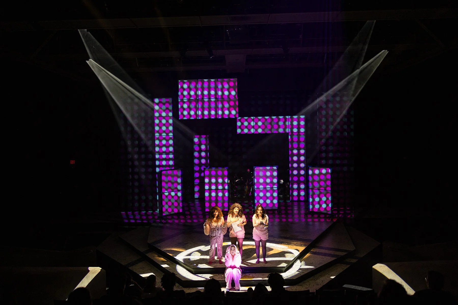 A stage with a geometric backdrop of lit panels displaying purple and pink dots. Four performers, all women, stand in front of the stage, one kneeling in front and three standing behind her, illuminated by spotlights.