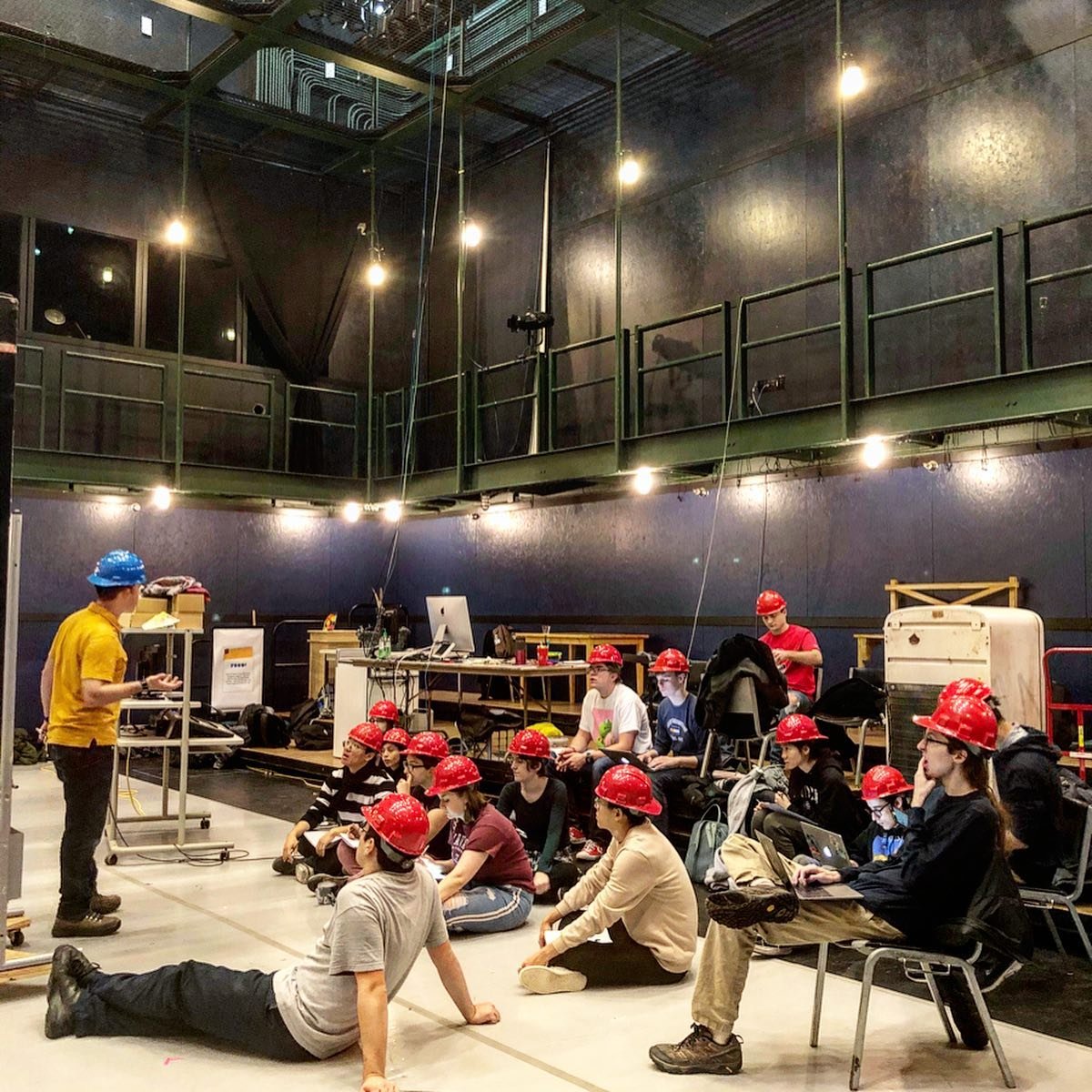 Group of people wearing red safety helmets, sitting and listening to a man in a blue helmet in an industrial-style room with black walls, yellow shirt, and computer equipment.