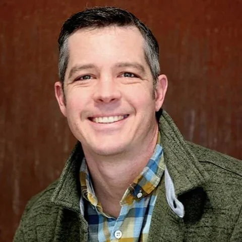 Portrait of a smiling man with short dark hair, wearing a green jacket over a plaid shirt, standing against a wooden background.
