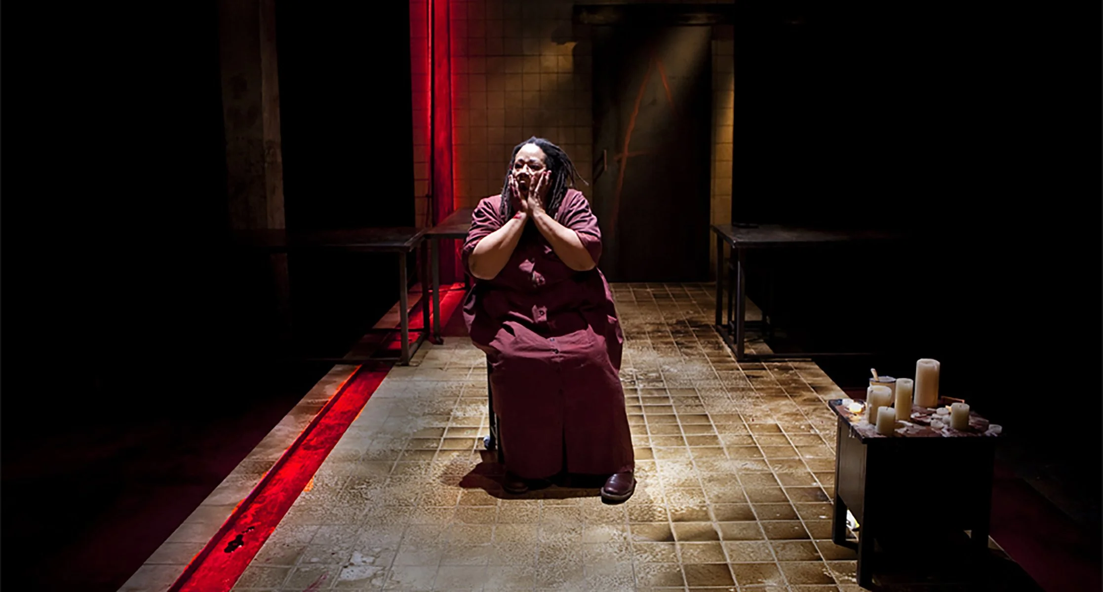 A woman with dreadlocks kneeling and holding her face in her hands in a dimly lit room with candles on a table in the foreground.