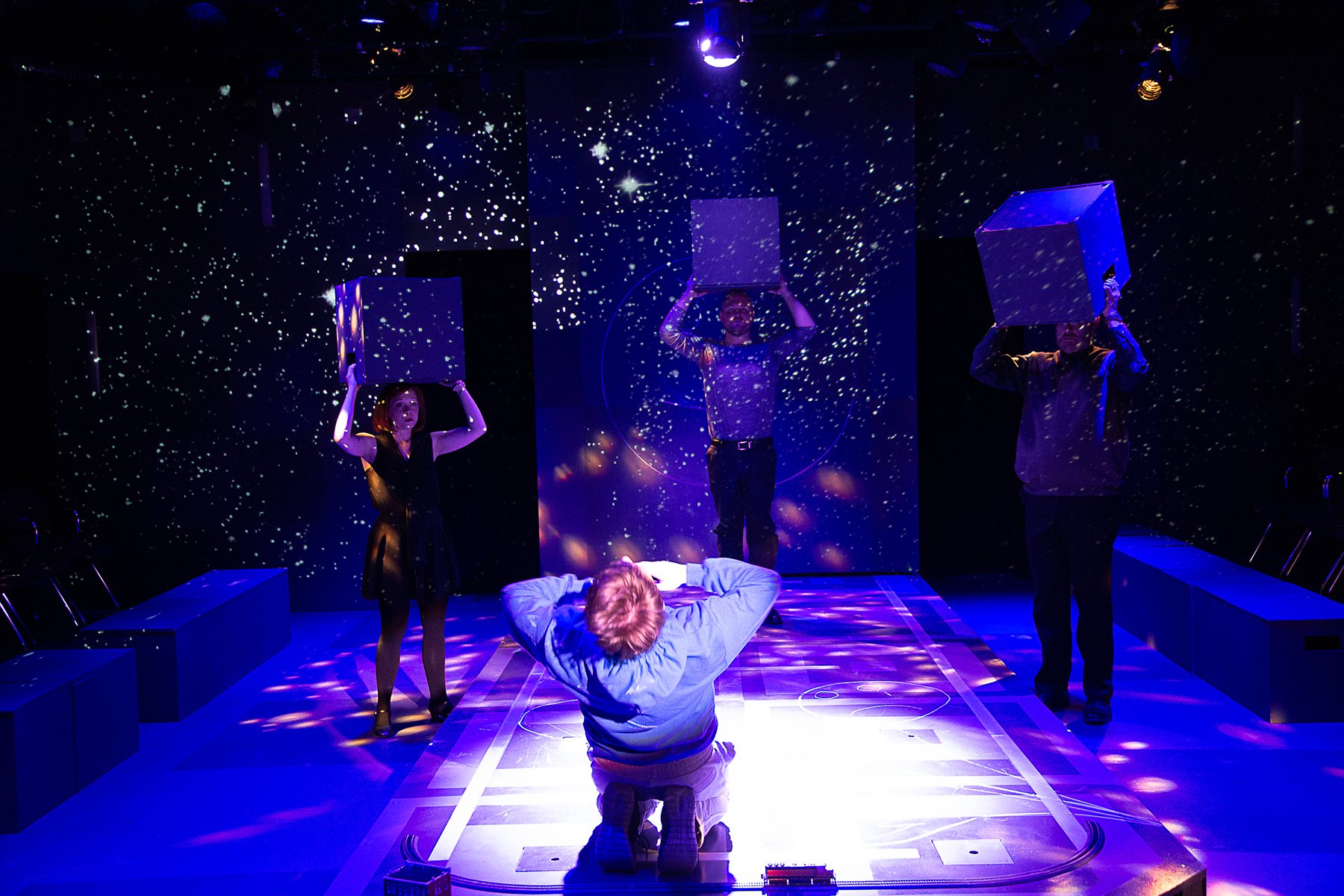 A theatrical scene with five people on stage surrounded by a starry background. Three are holding large cubes above their heads, while another is lying face down on the floor, and one is kneeling in front of them, with their back to the camera, takin