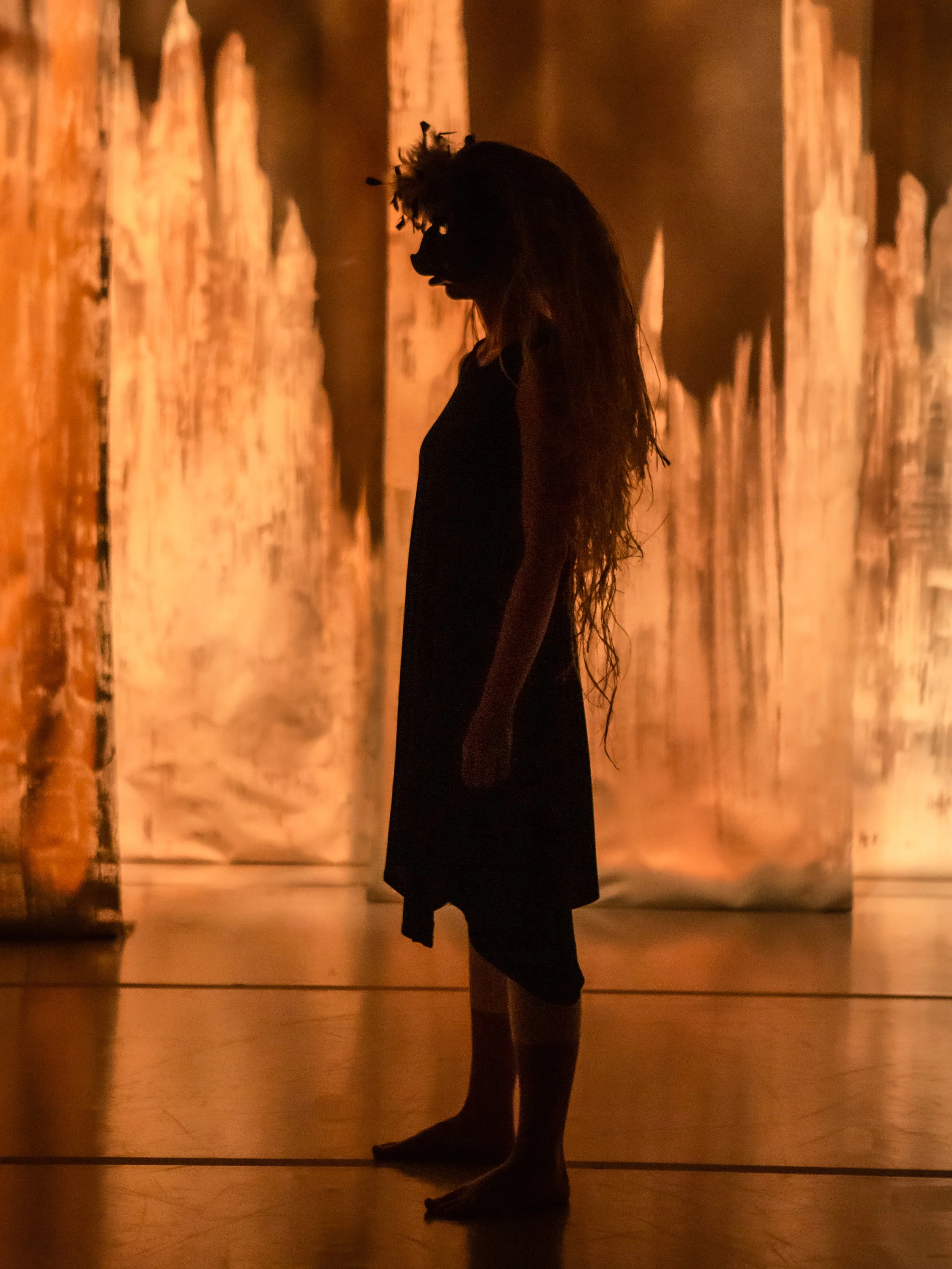 Silhouette of a woman standing in front of illuminated wooden background, with her head bowed and hair falling over her face.