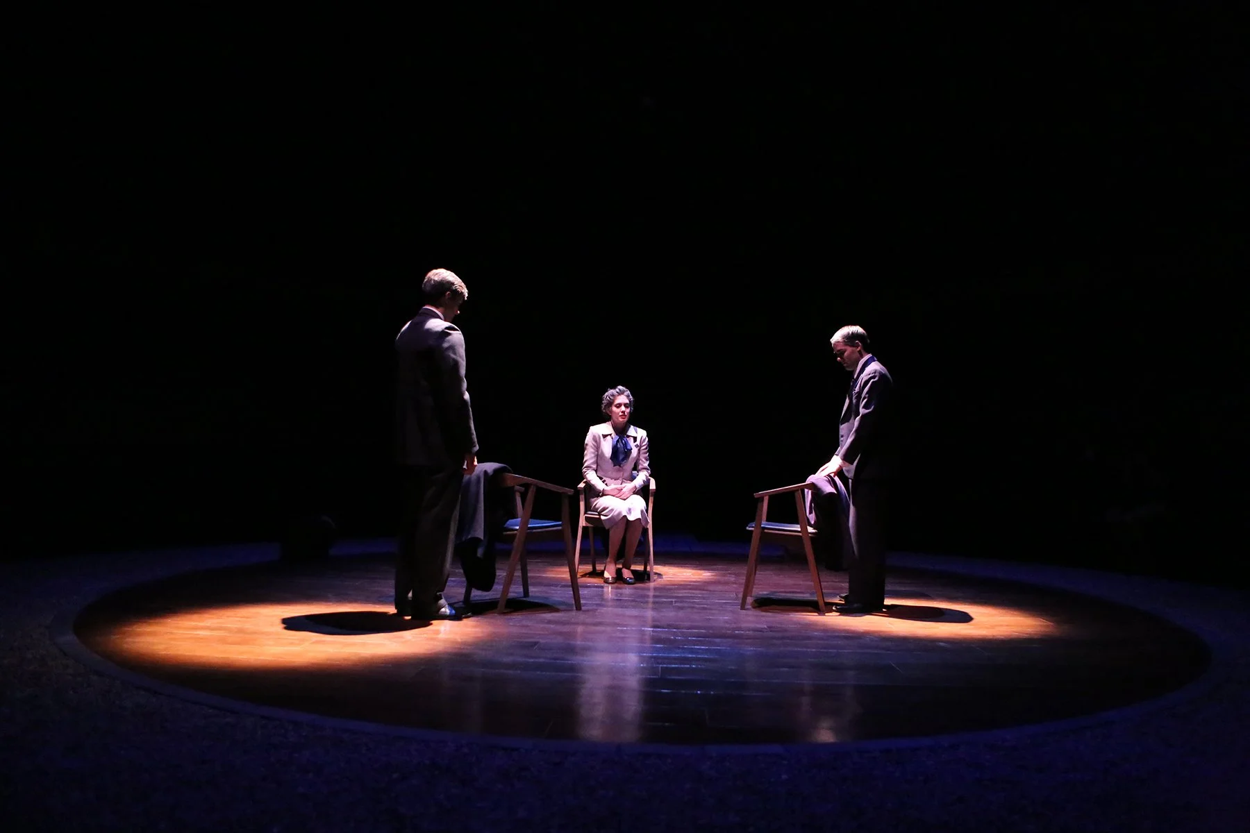 Three actors on a dark stage with spotlight, two standing and one sitting, portraying a scene in a theater or play.