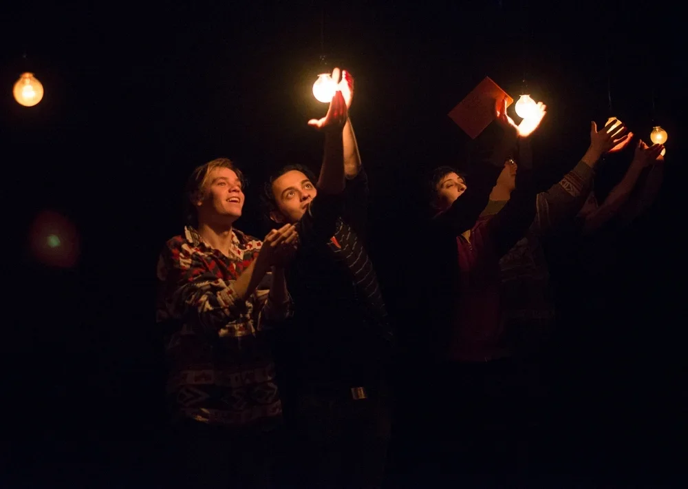 Three people holding glowing light bulbs and one person holding a red book, standing in a dark room with hanging light bulbs.