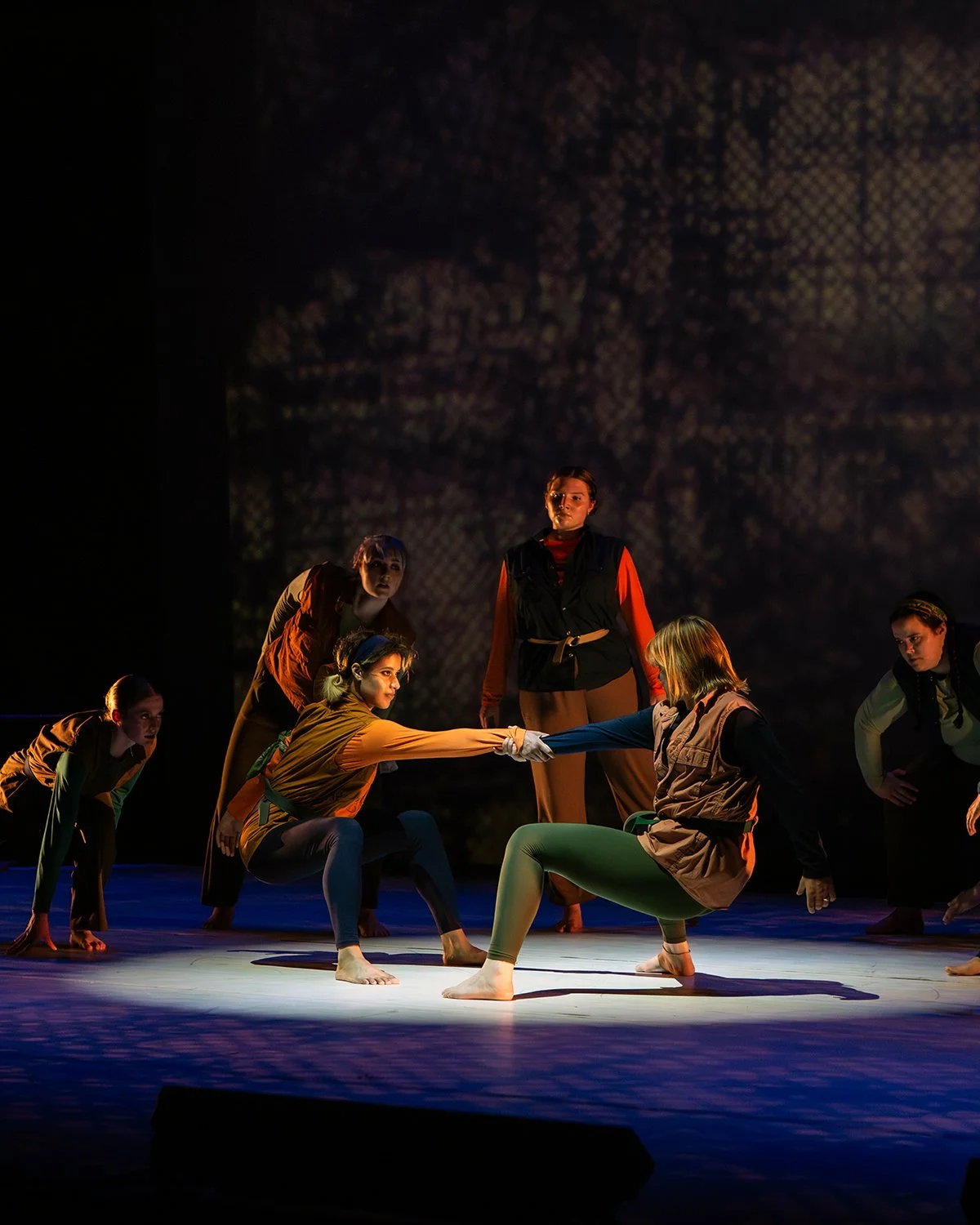 A theatrical performance with six actors on stage, some kneeling and others standing, engaging in a dramatic scene with a dark, textured backdrop.