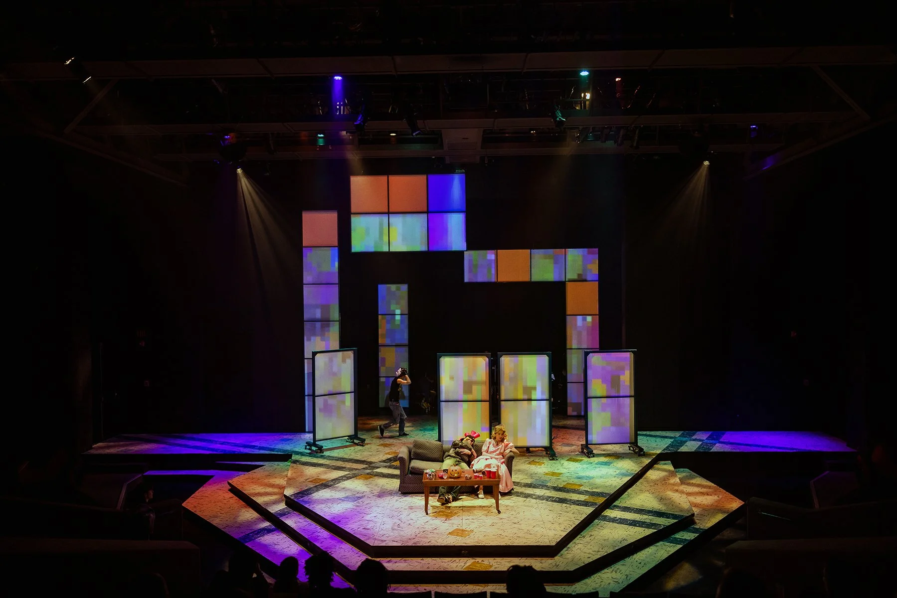 Stage with colorful digital screens forming the shape of a house, lit by stage lights, with performers and children seated in front.