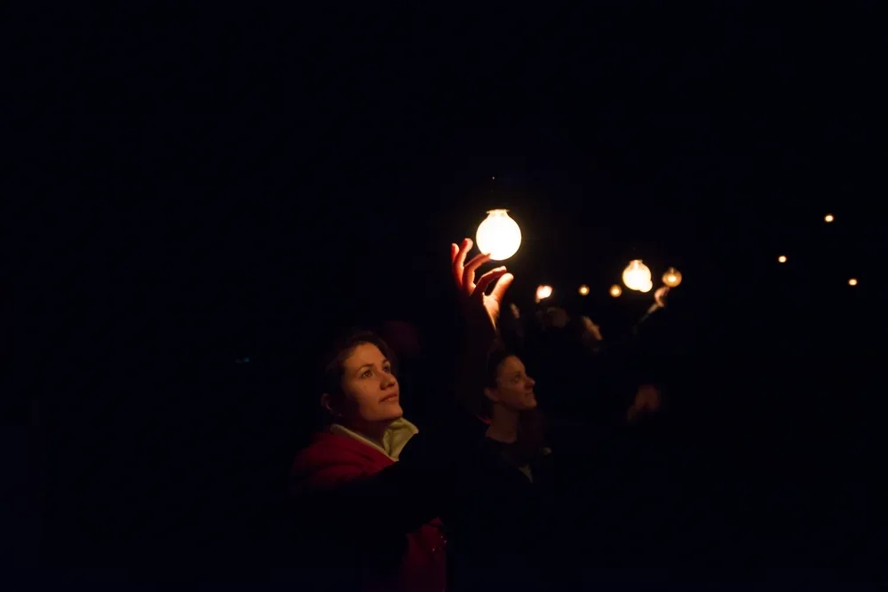 People holding lanterns at night, with one woman in the foreground reaching out to touch a glowing lantern.