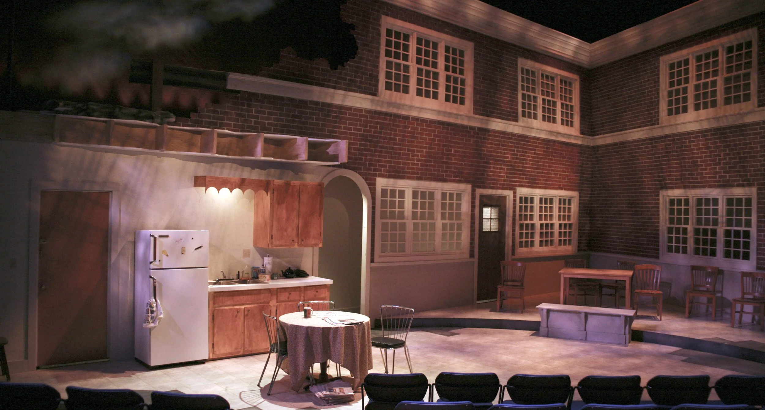 Interior of a theater stage set resembling a brick building with multiple windows, a kitchen area with wooden cabinets, a table with chairs, and a small deck area.
