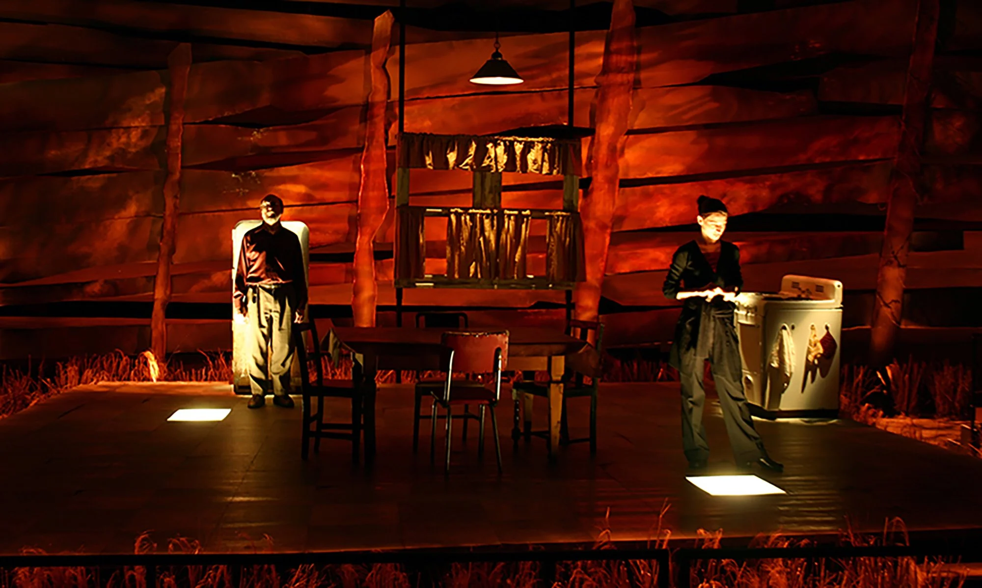 Stage scene from a theatrical production with two actors, one man in a shirt and trousers standing near a chair, and a woman in dark clothing working with a laundry basket. The stage has a large wooden log backdrop and minimal furniture, illuminated 