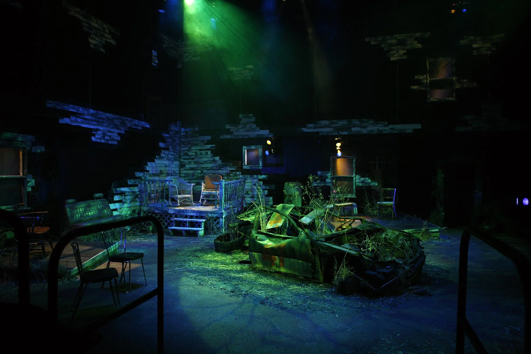 Theater stage set designed to look like an outdoor scene with an overgrown garden, broken car, and worn furniture, illuminated with green, blue, and purple lights.