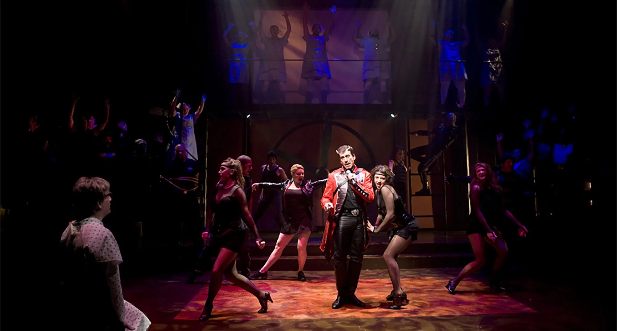 The image depicts a lively stage performance with multiple performers. In the foreground, a man dressed in a red and black jacket stands center stage, surrounded by women in black outfits dancing. Additional performers are on stairs and a balcony in 
