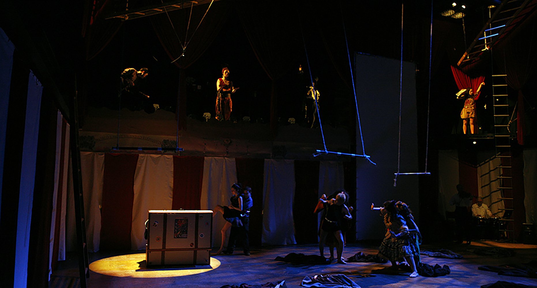 The image shows a theatrical stage with performers preparing for a show, including some on a balcony and others on the stage floor, with dark lighting and various props and costumes.