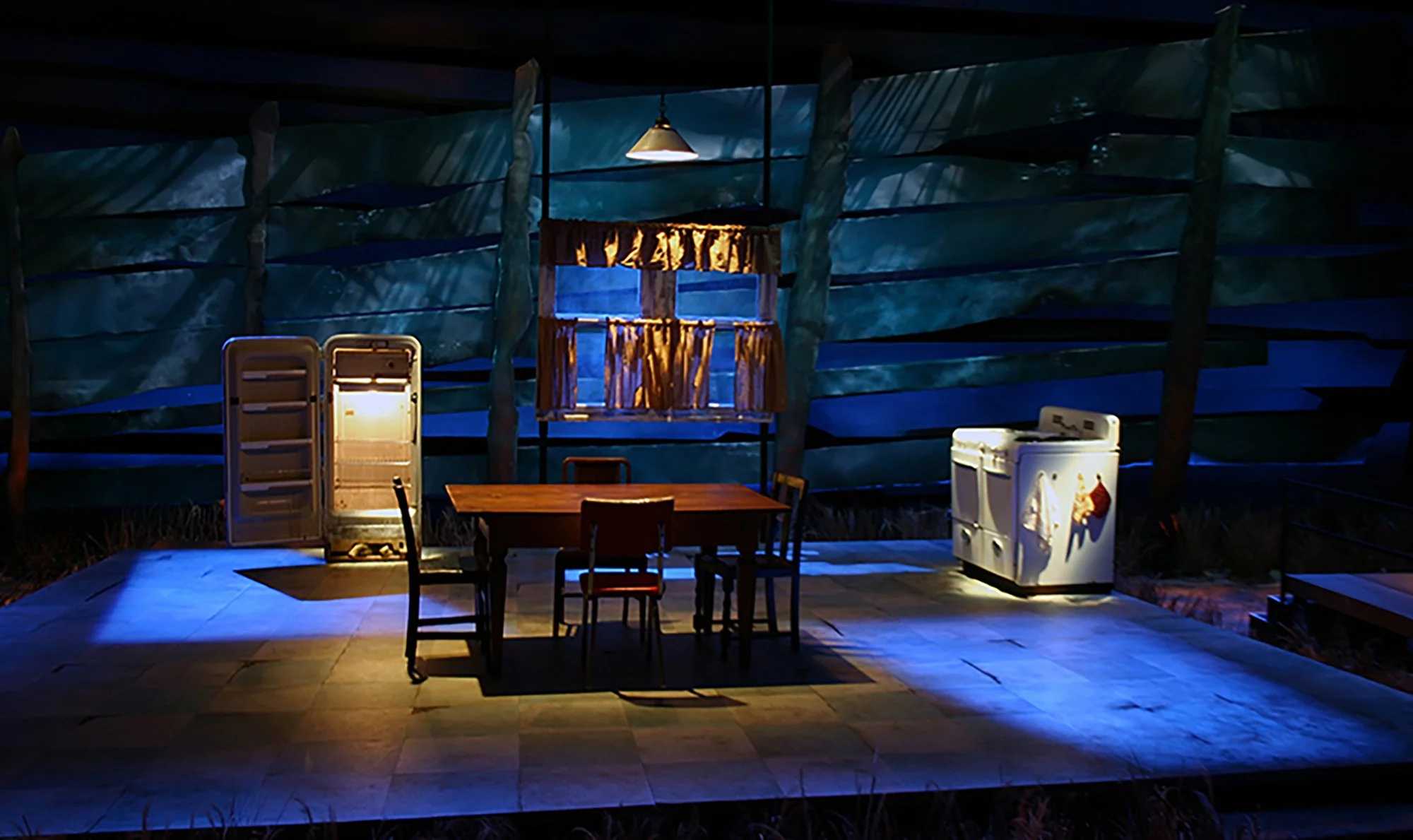 A dimly lit outdoor dining area with a wooden table, chairs, a small refrigerator, and a cake with lit candles, surrounded by a blue fabric backdrop.