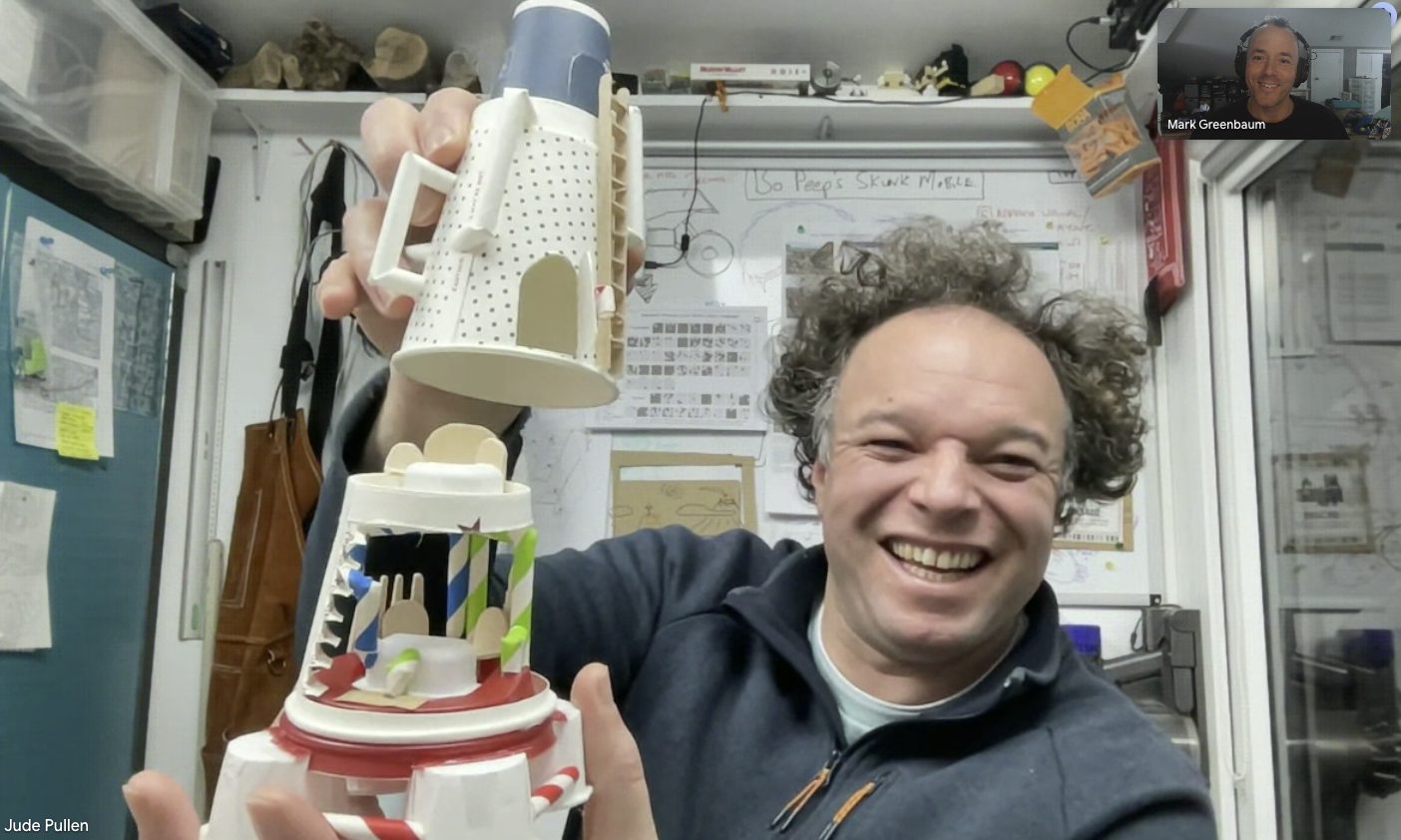 Man with curly hair smiling and holding a colorful model made of recycled materials in an office or workshop setting.