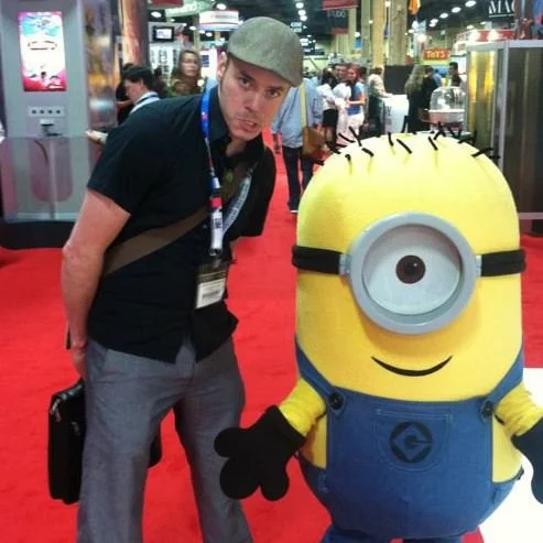 A man standing next to a life-sized Minions character at a convention or expo, with booths and other attendees in the background.