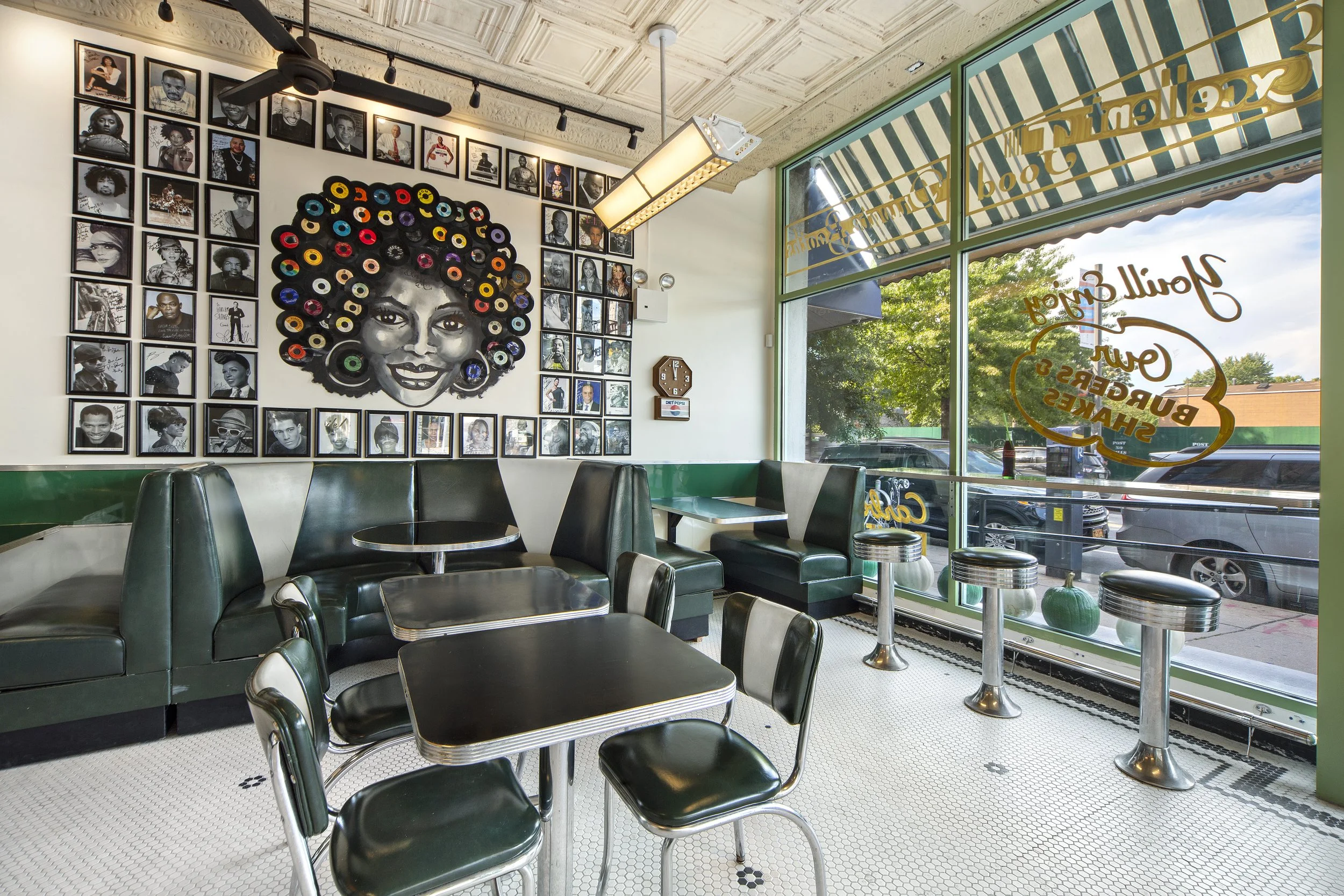 Interior of a vintage-style diner with green booths and diner stools, decorated with black-and-white framed photographs of various people on the wall, including a large artistic mural of a smiling woman made of vinyl records, and a window with the words 'Gourmet' and 'Burgers' etched on the glass, showing trees and parked cars outside.