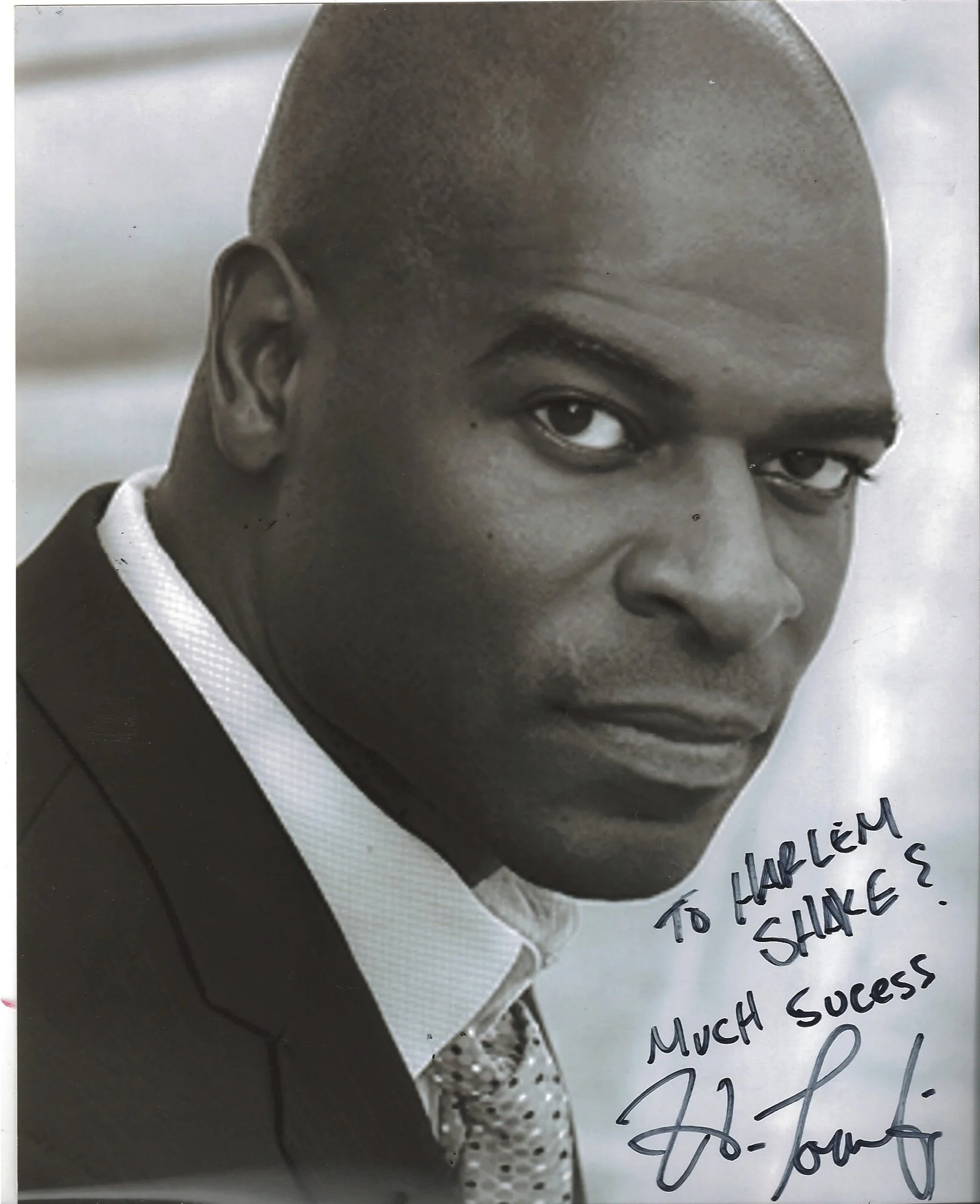 A black-and-white photograph of a man dressed in a suit with a tie, with handwritten text on the right side of the image that reads 'To Harold, Much success,'. The man's face is partially visible, showing the right eye, forehead, and part of his mout