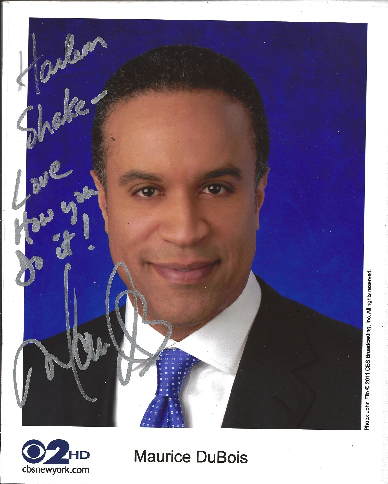 Close-up of Maurice DuBois, an African American man, wearing a black suit, white shirt, and blue tie with white checkered pattern, against a blue background. The image appears to be from a professional event or interview, with his name displayed at t