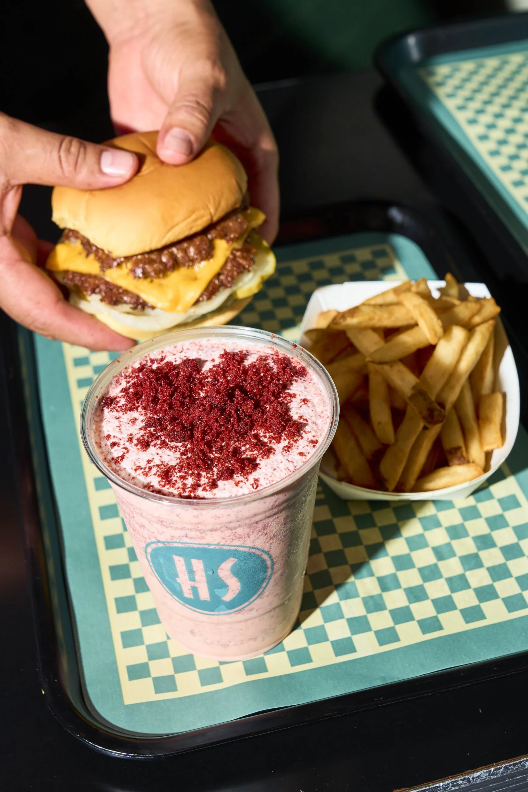 A burger with beef patty, cheese, and bun being assembled on a tray, with a side of French fries and a strawberry flavored milkshake with red sprinkles on top.