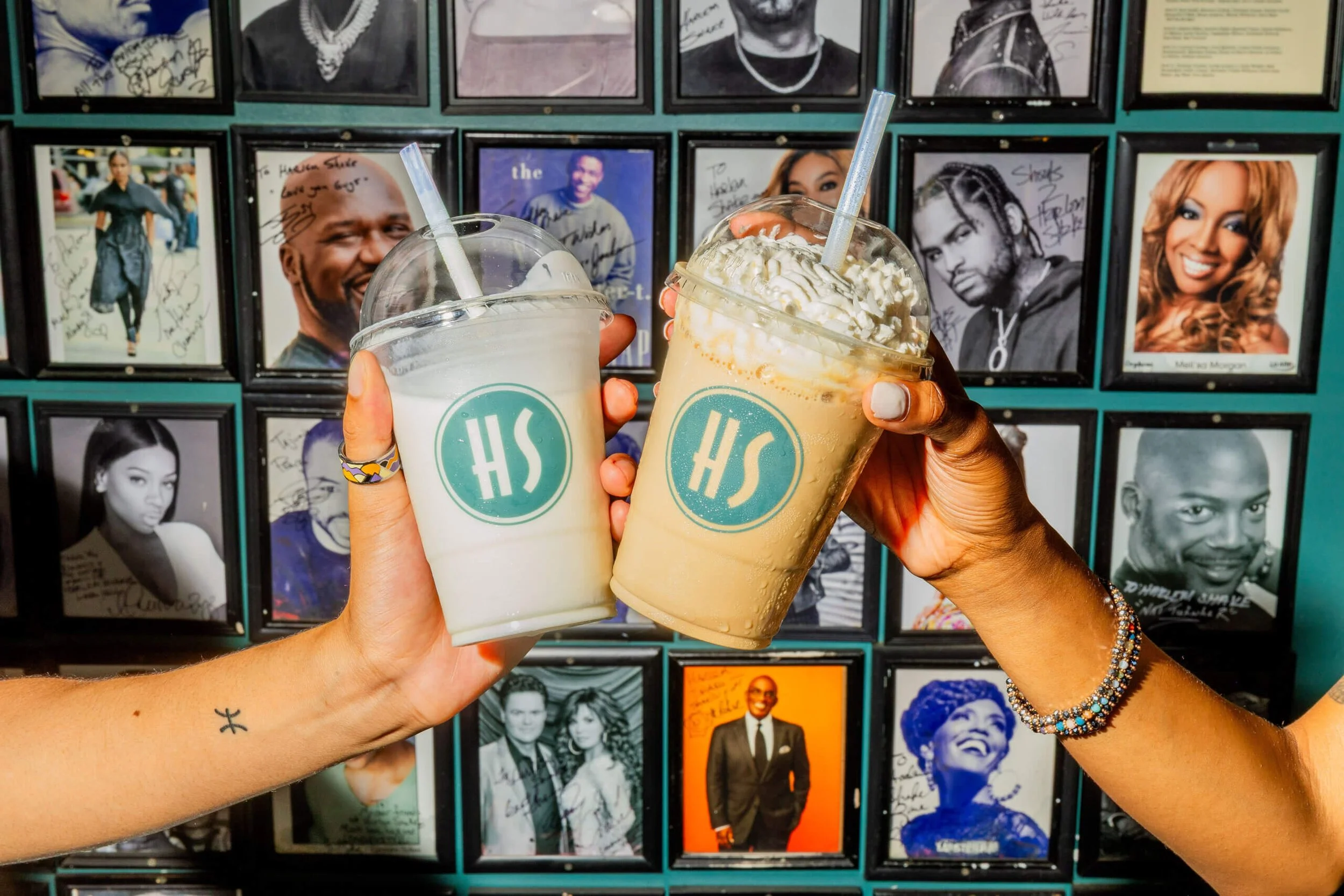 Two hands holding drinks with whipped cream, one beverage is a milkshake and the other is a coffee drink, both with clear cups and straws, set against a background of framed black and white photographs of various celebrities and public figures.