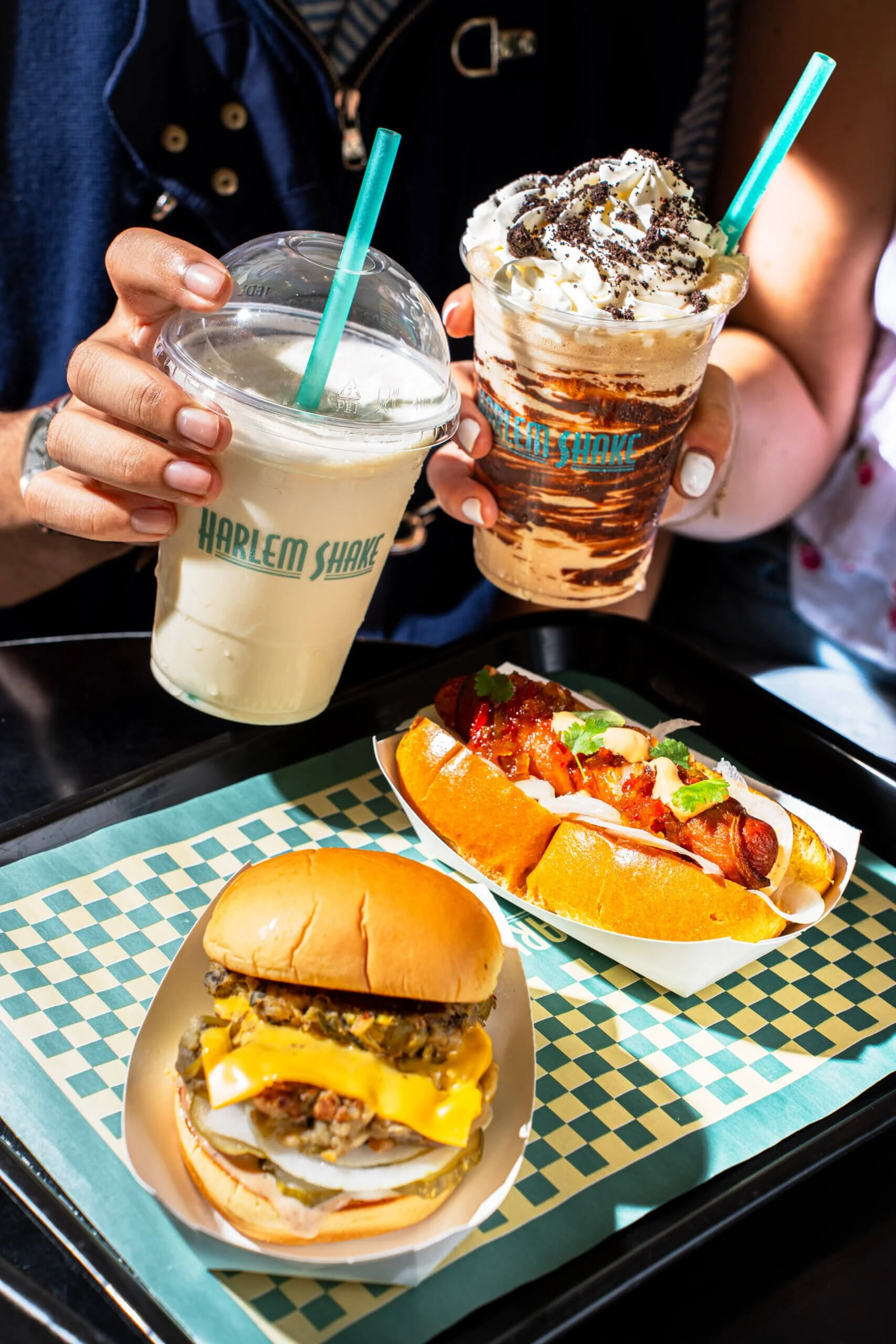 Two people holding milkshakes, one vanilla with a green straw and whipped cream, the other chocolate with whipped cream and cookie crumbs. A tray with a cheeseburger, hot dog with toppings, and a bun is on the table in front.