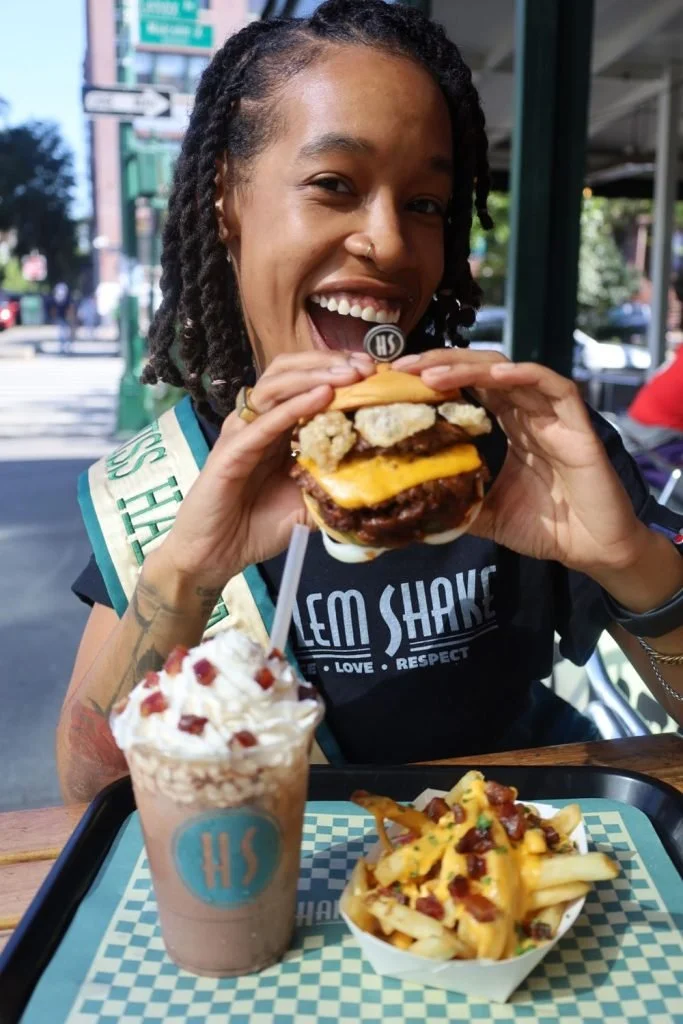 Woman holding a large hamburger with fries and a milkshake on the table in front of her at an outdoor dining area.