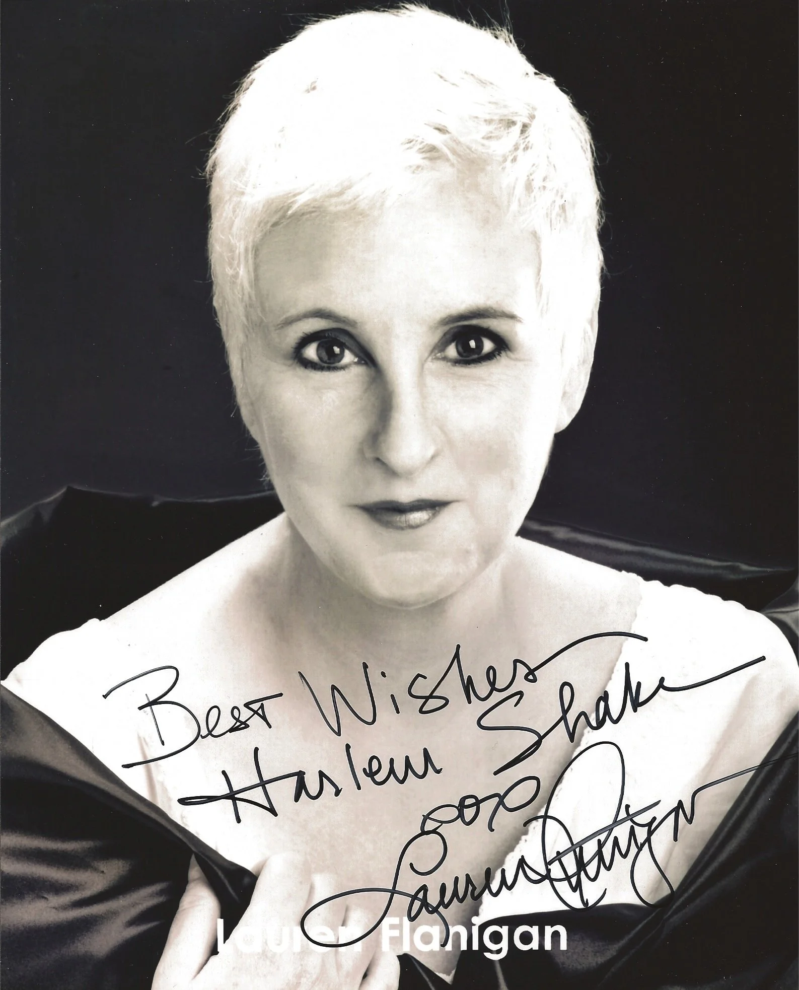 Black and white photograph of a woman with short, curly, platinum blonde hair, wearing a white top and a black jacket, holding a whiteboard with handwritten message, and looking directly at the camera.