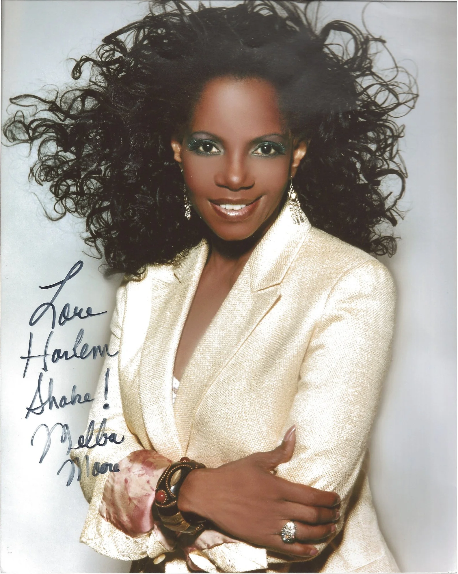 Close-up portrait of an African American woman with curly hair, wearing a gold blazer, earrings, and rings, smiling with her arms crossed, against a plain background.