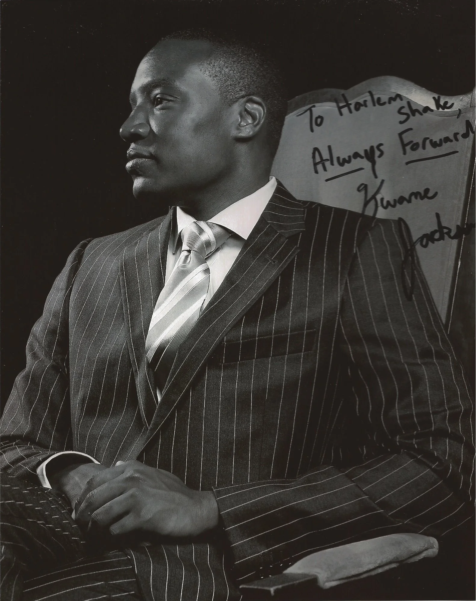 A black and white photograph of a man in a checkered suit with a striped tie, sitting with his hand on his lap. There is a note on the backrest of his chair that reads, "To Harlem Shake Always Forward Kwame."