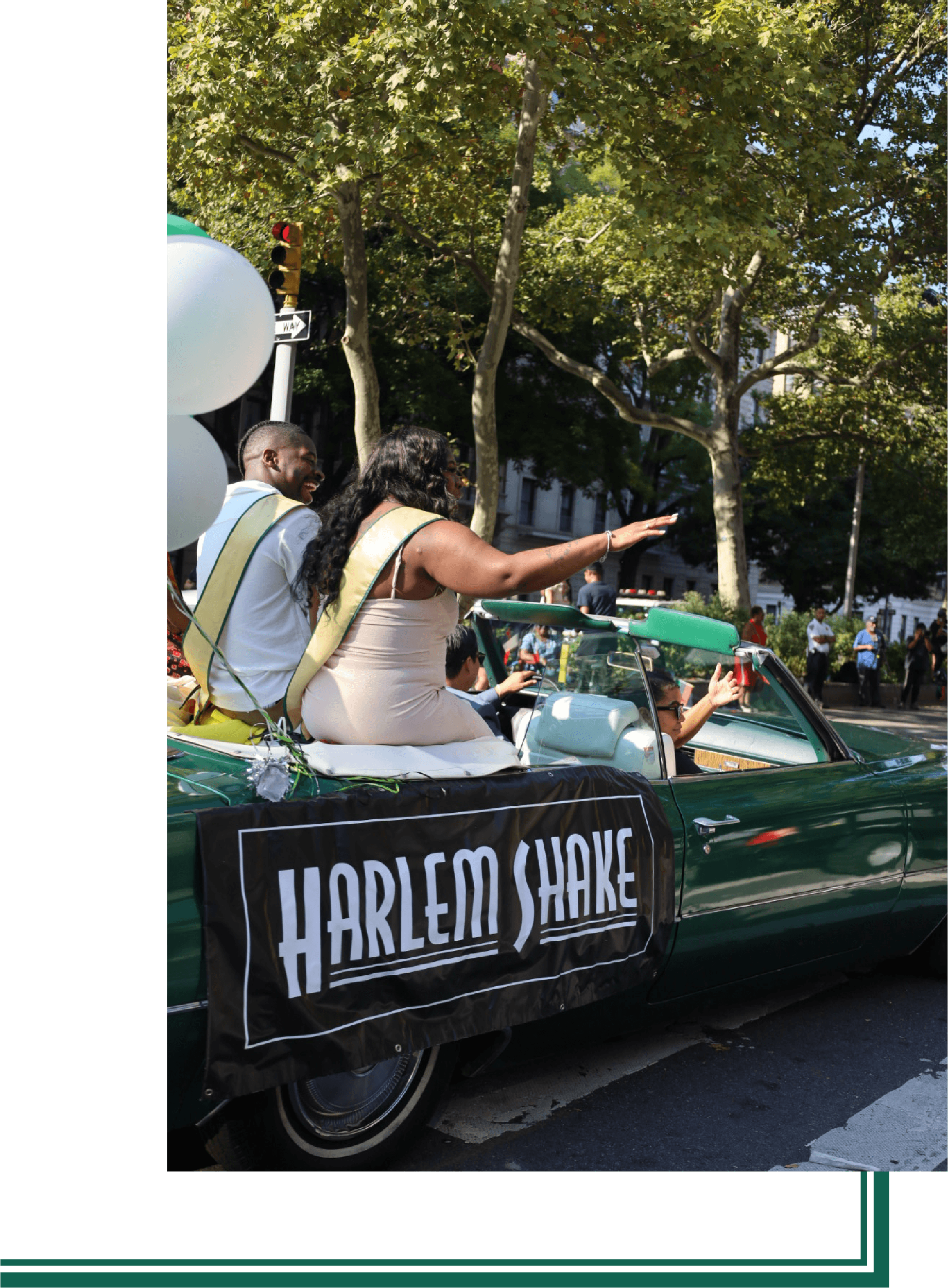 A parade float decorated with white balloons and a banner that reads "Harlem Shake". Two people are sitting on top of the float, one man and one woman, waving and smiling. The float is on a city street with trees and buildings in the background.