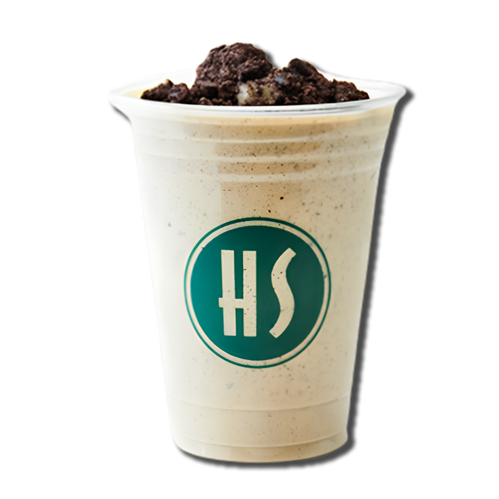 A creamy milkshake topped with crushed Oreo cookies in a clear plastic cup with a green circular logo that has the letters 'HS'.