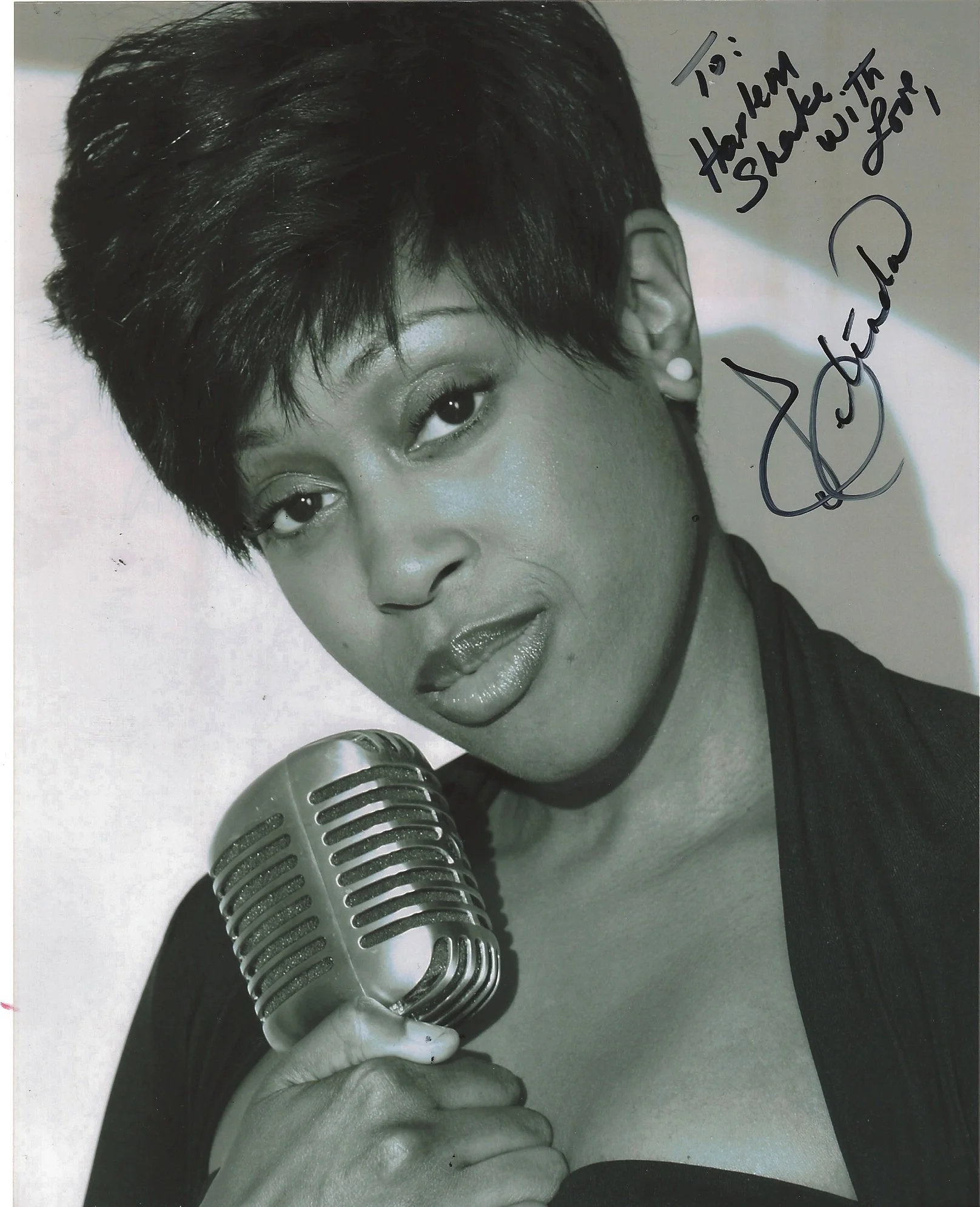 Black and white photo of a woman with short hair singing into a vintage microphone. She has eye makeup, earrings, and shiny lipstick, with a signature and handwritten message in the upper right corner.