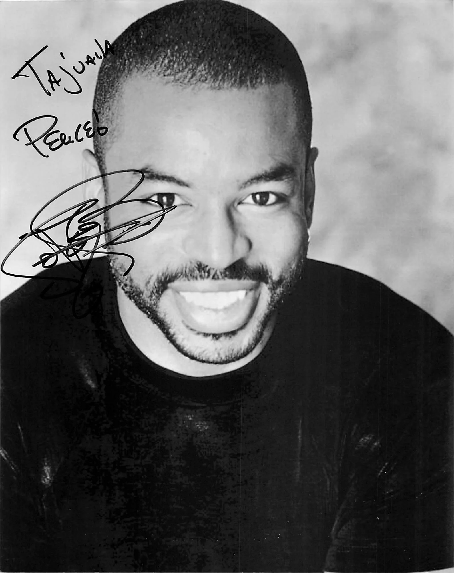 A black-and-white portrait of a young man with a close-cropped haircut, smiling widely showing teeth, with handwritten text and a signature on the left side of the image.