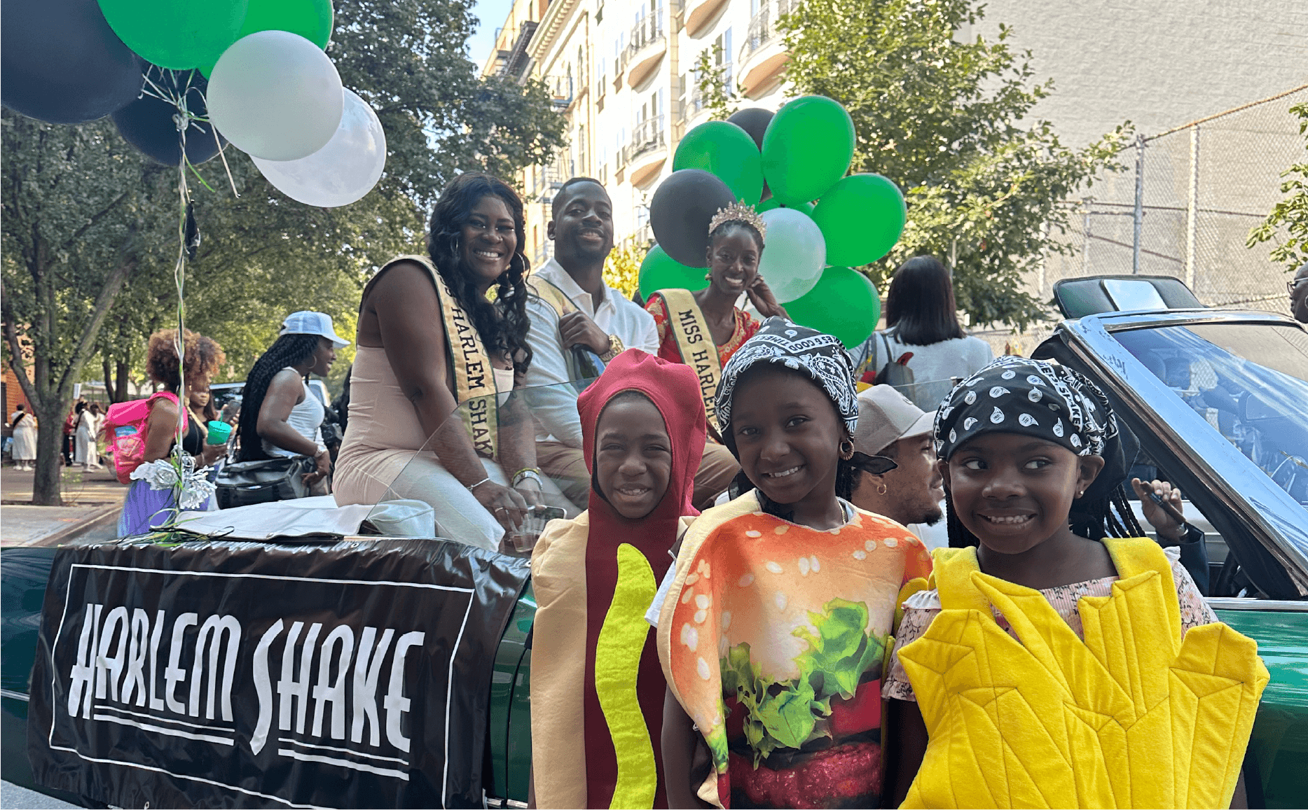 A parade float with participants, including women wearing sashes that say 'Miss Harlem Shake,' and children dressed in colorful costumes, surrounded by green, white, and black balloons, with a city street and apartment buildings in the background.