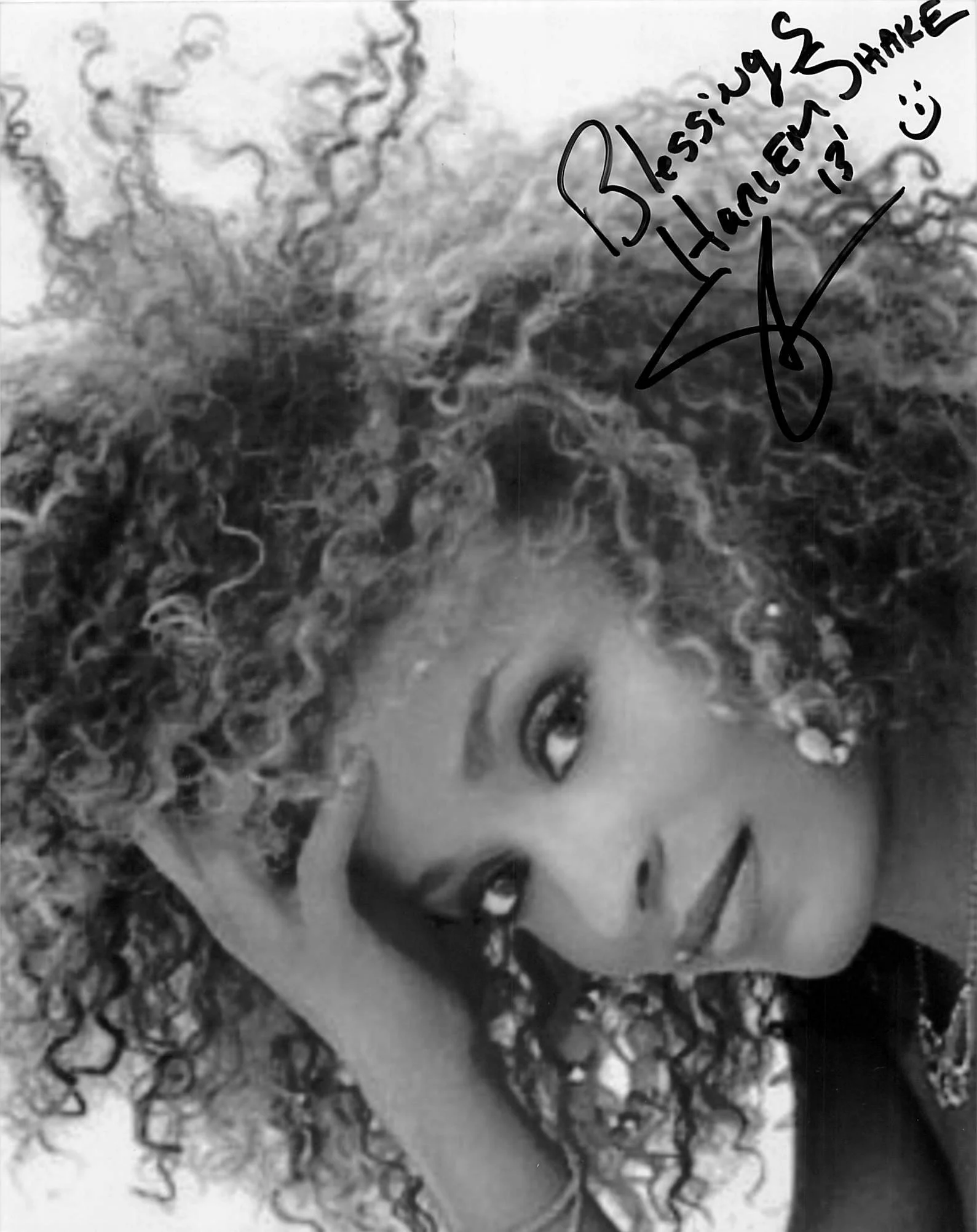 A black and white drawing of a smiling woman's face with curly hair. There is a handwritten message on the right side that says, 'Blessings & Happiness :)' with a smiley face, and the signature appears to be 'X'.