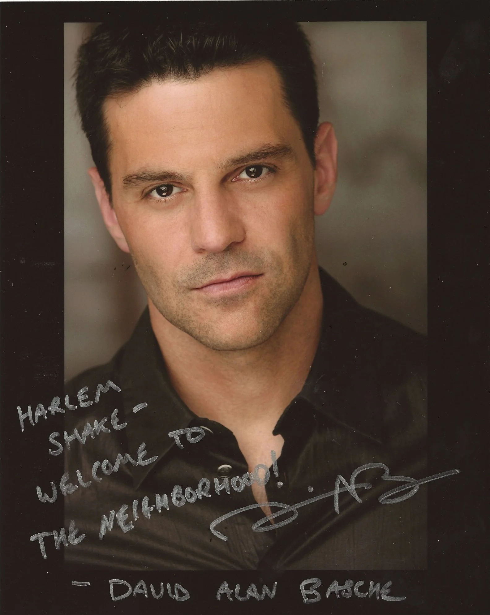A portrait of a man with dark hair wearing a black shirt, smiling softly, with handwritten text in silver on the photograph that reads, 'WELCOME HARLEM SHAWK, TO THE NEIGHBORHOOD! DAVID ALAN BASCHE'.