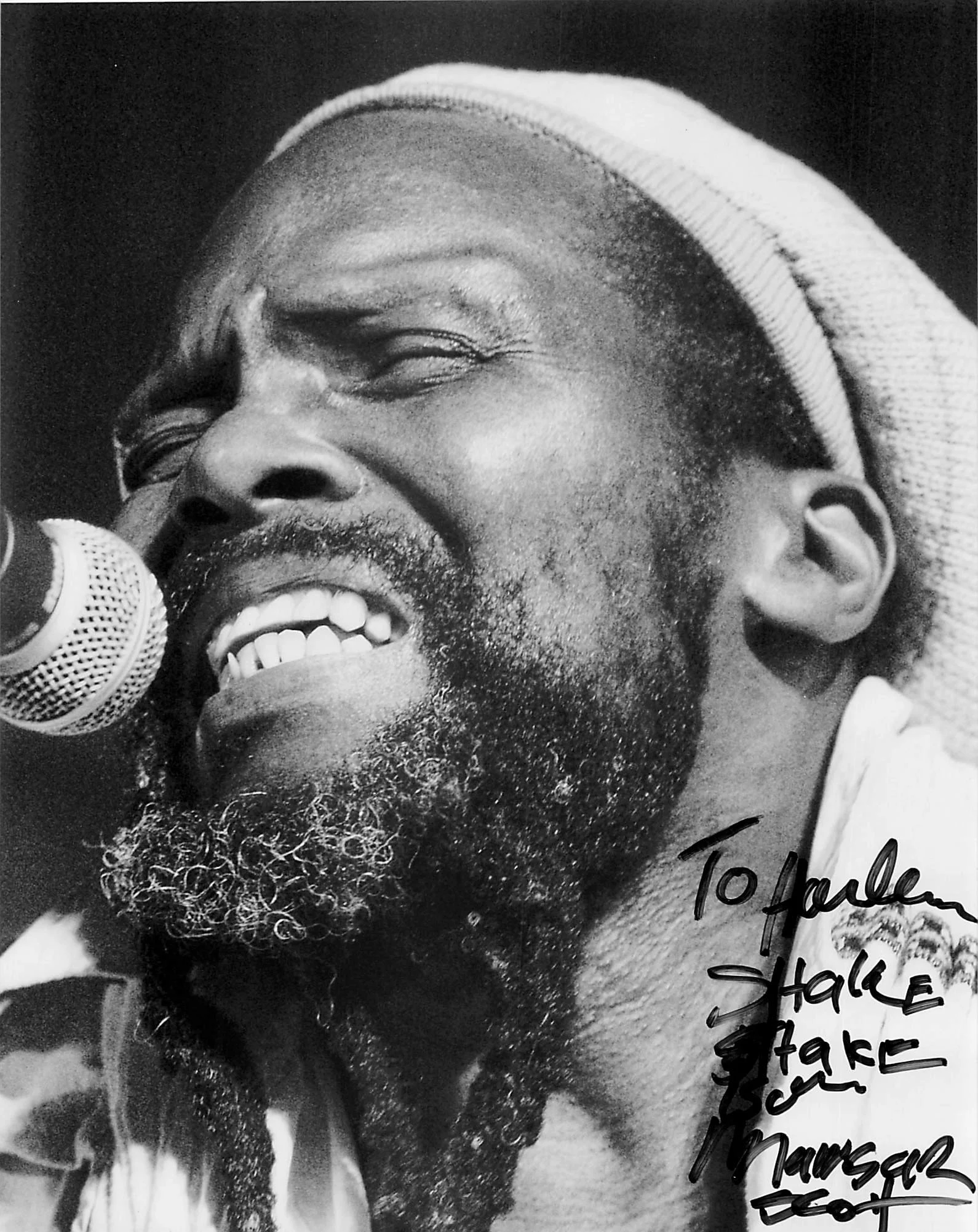 A black and white photo of a man singing into a microphone, wearing a hat and smiling. There is handwritten text on the bottom right corner of the photo.