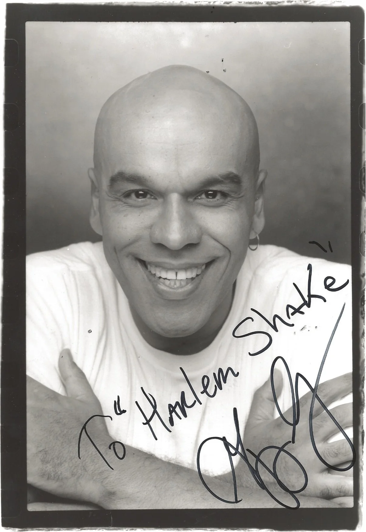 Black and white photograph of a smiling bald man with earrings, wearing a light-colored shirt, with handwritten text that says, "To Harlem Shake" and a signature on the bottom right.