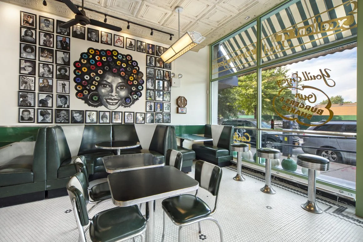 Interior of a retro-style cafe with black and white photos on the wall, a TV, and a colorful portrait of a woman with an afro made of records. There are black and green booths, black tables, and black chairs near large window with yellow and green striped awning outside.