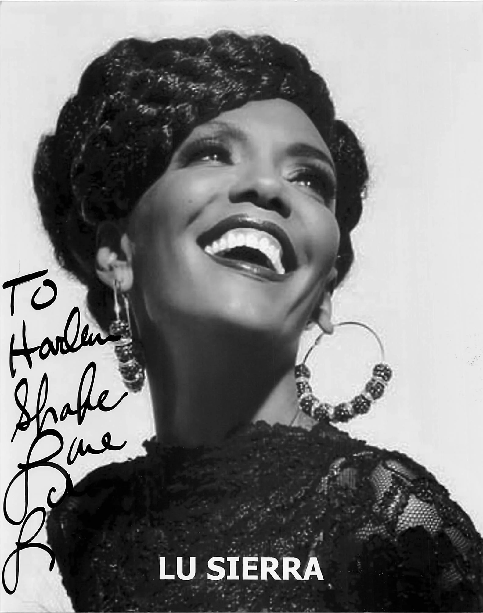 Black and white photo of a woman with short, textured hair and a wide smile, wearing large earrings and a beaded necklace, with handwritten-style text that reads 'To Hardene'.