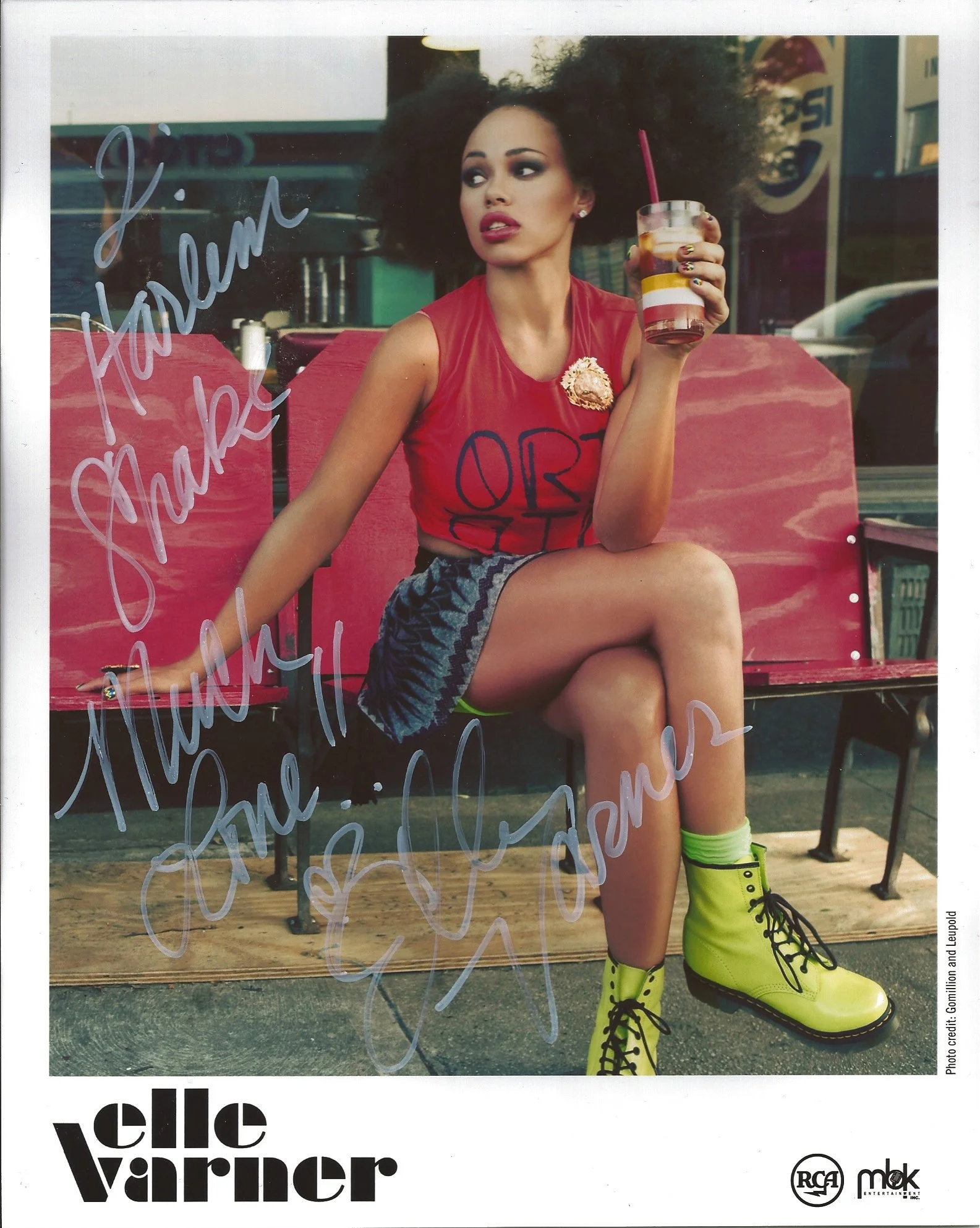 A woman with big curly hair sitting on a pink bench outside a café, holding a layered drink with a pink straw. She wears a red sleeveless top, a patterned skirt, neon yellow boots, and has colorful nail polish. There are handwritten messages on the g