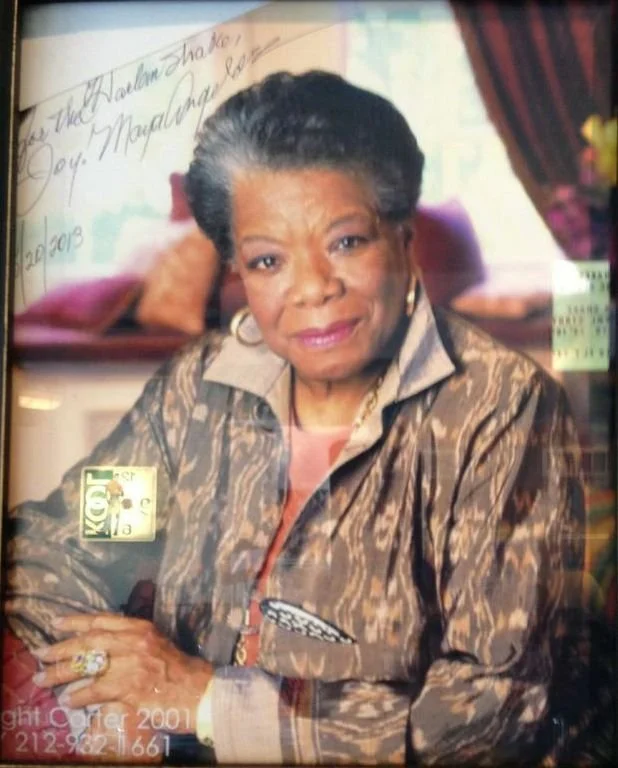 Portrait of an older woman with gray hair, wearing a patterned jacket, seated with arms crossed, indoors with a window and curtains in the background.