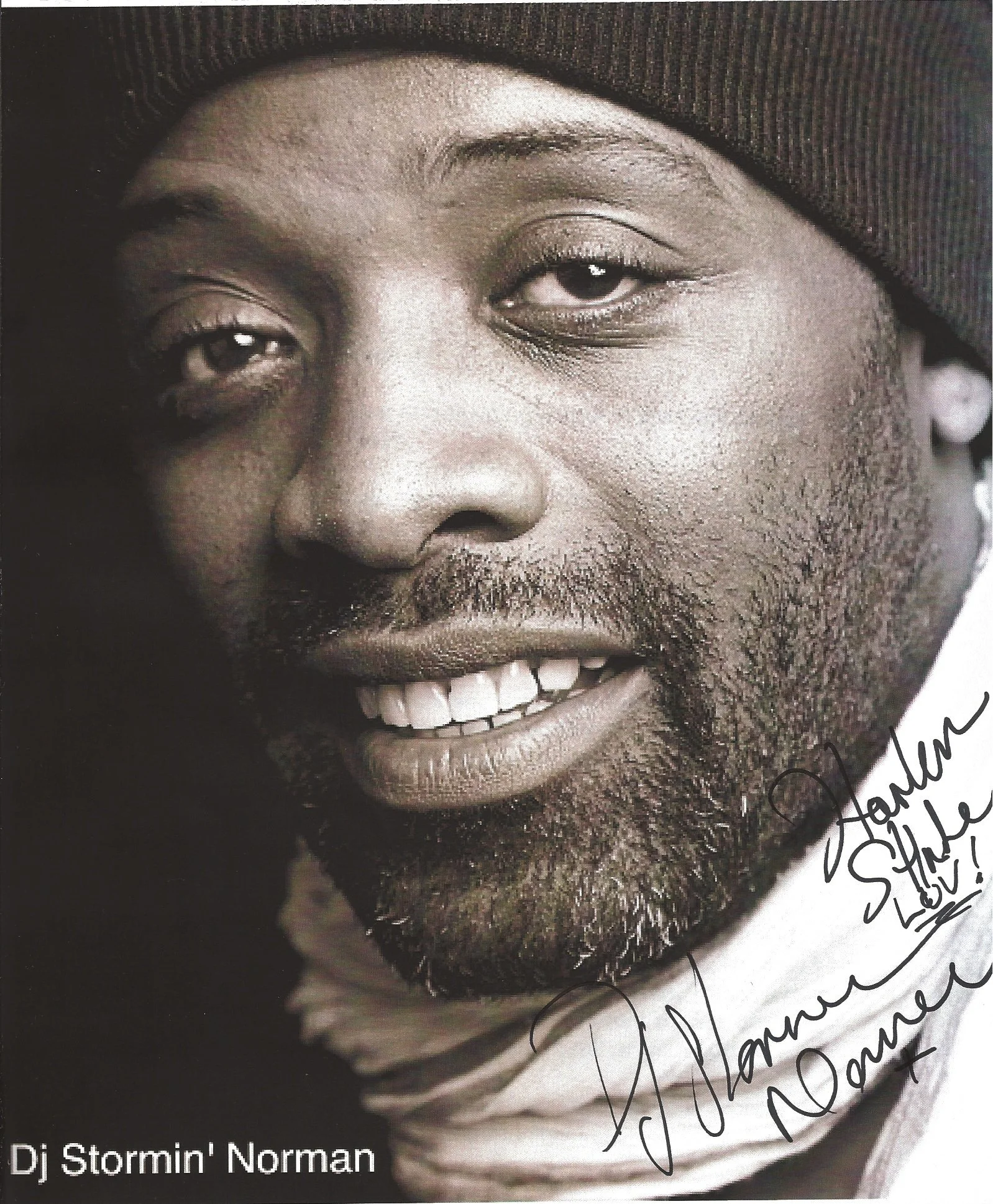 Close-up black and white photograph of a smiling man wearing a beanie and a white hoodie with handwritten signatures on the bottom right corner.