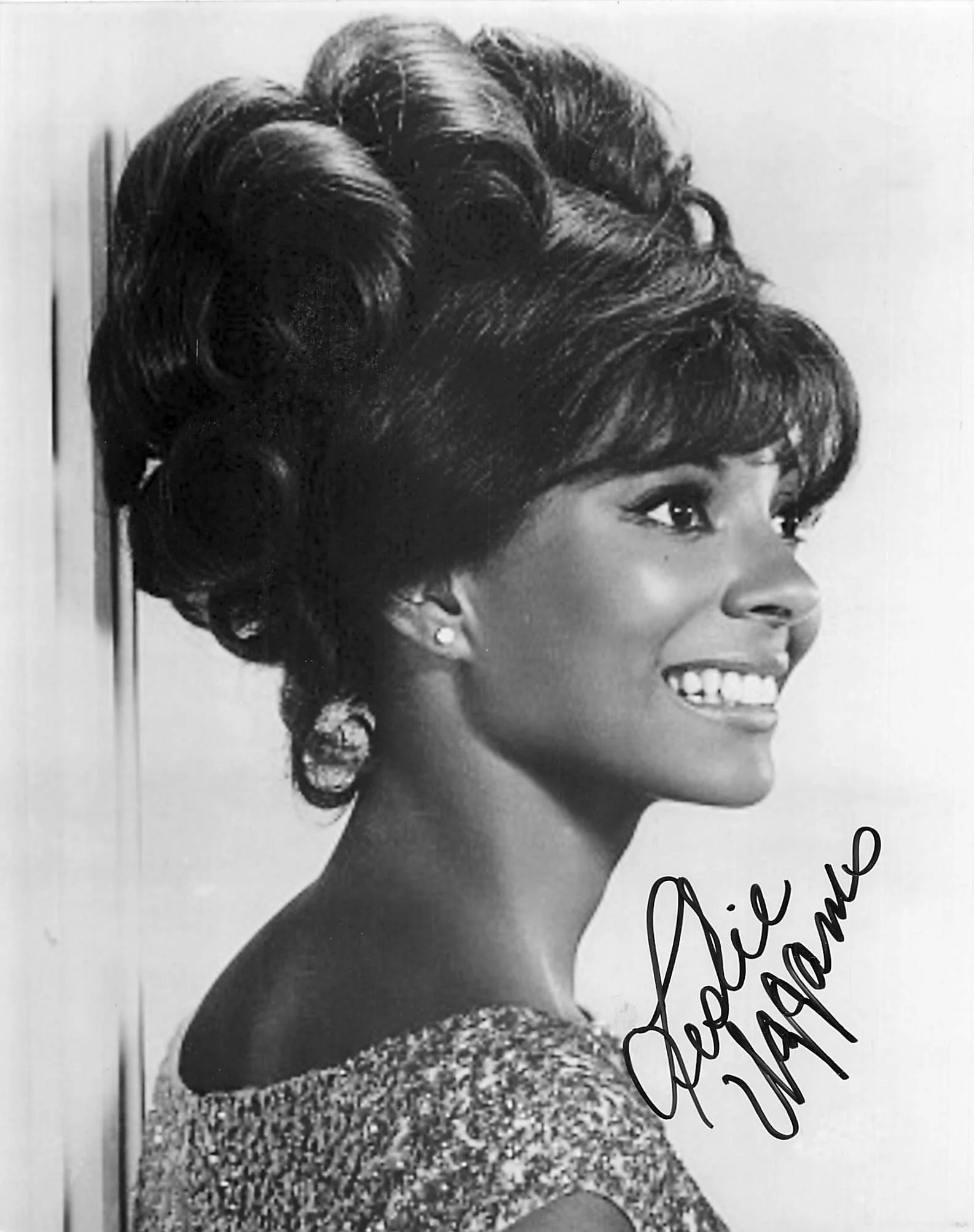A black-and-white photograph of a smiling woman with short, styled hair, wearing earrings, and dressed in an elegant outfit with a textured, shiny neckline. The image appears to be a vintage portrait.